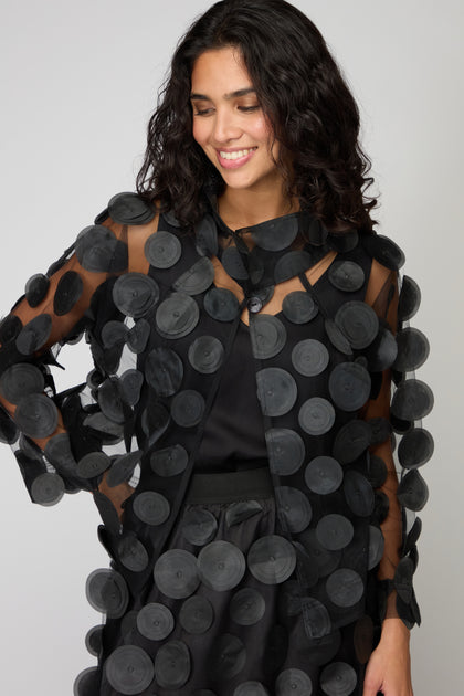 Smiling woman in the Cut Out Circles Jacket—a sheer black mesh piece with bold circular textures—looks down against a plain background, giving off a chic Scandinavian-inspired vibe.