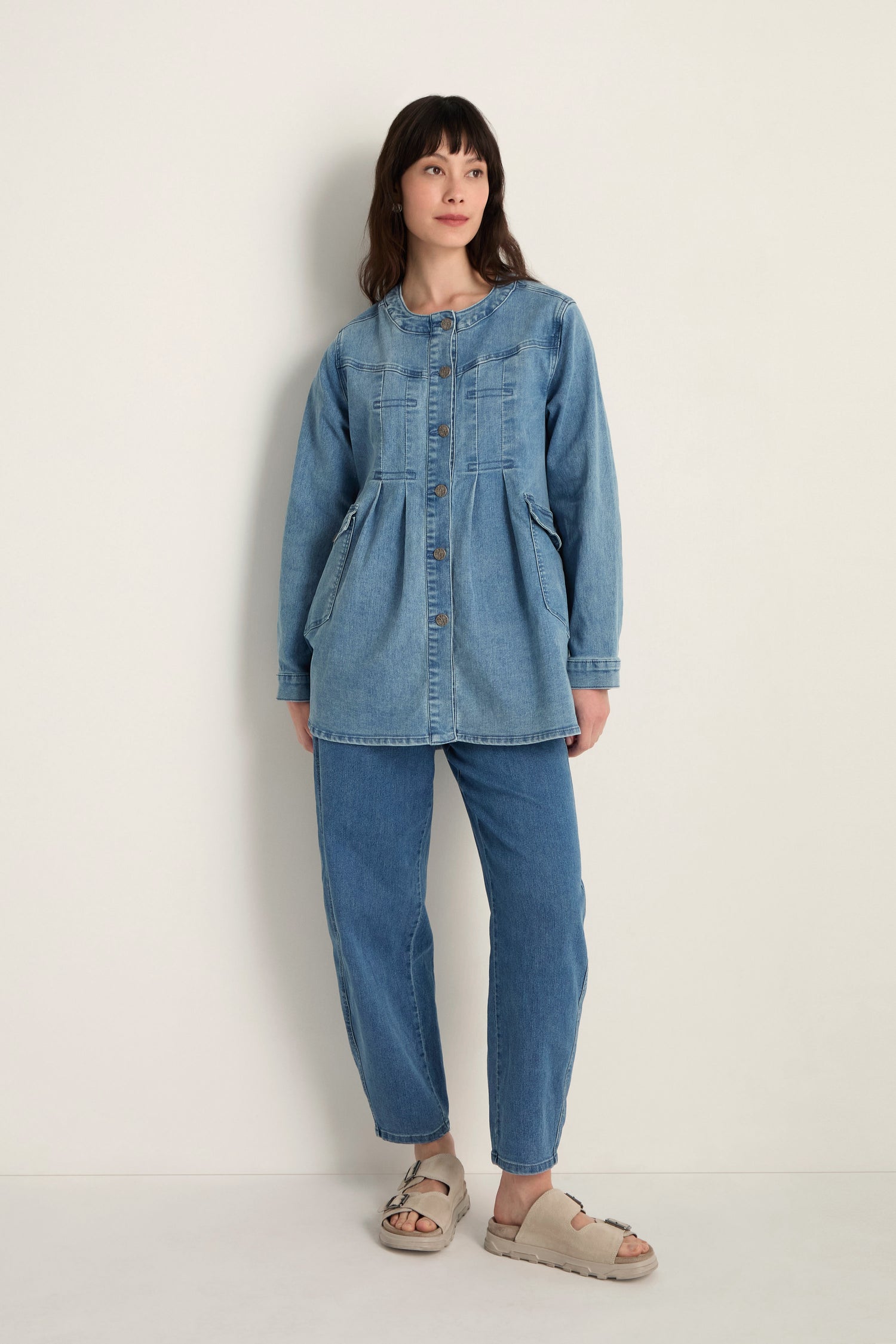 A woman stands against a plain wall wearing the Long Denim Jacket, a modern layering piece made from soft stretch denim, styled with blue jeans and beige sandals.