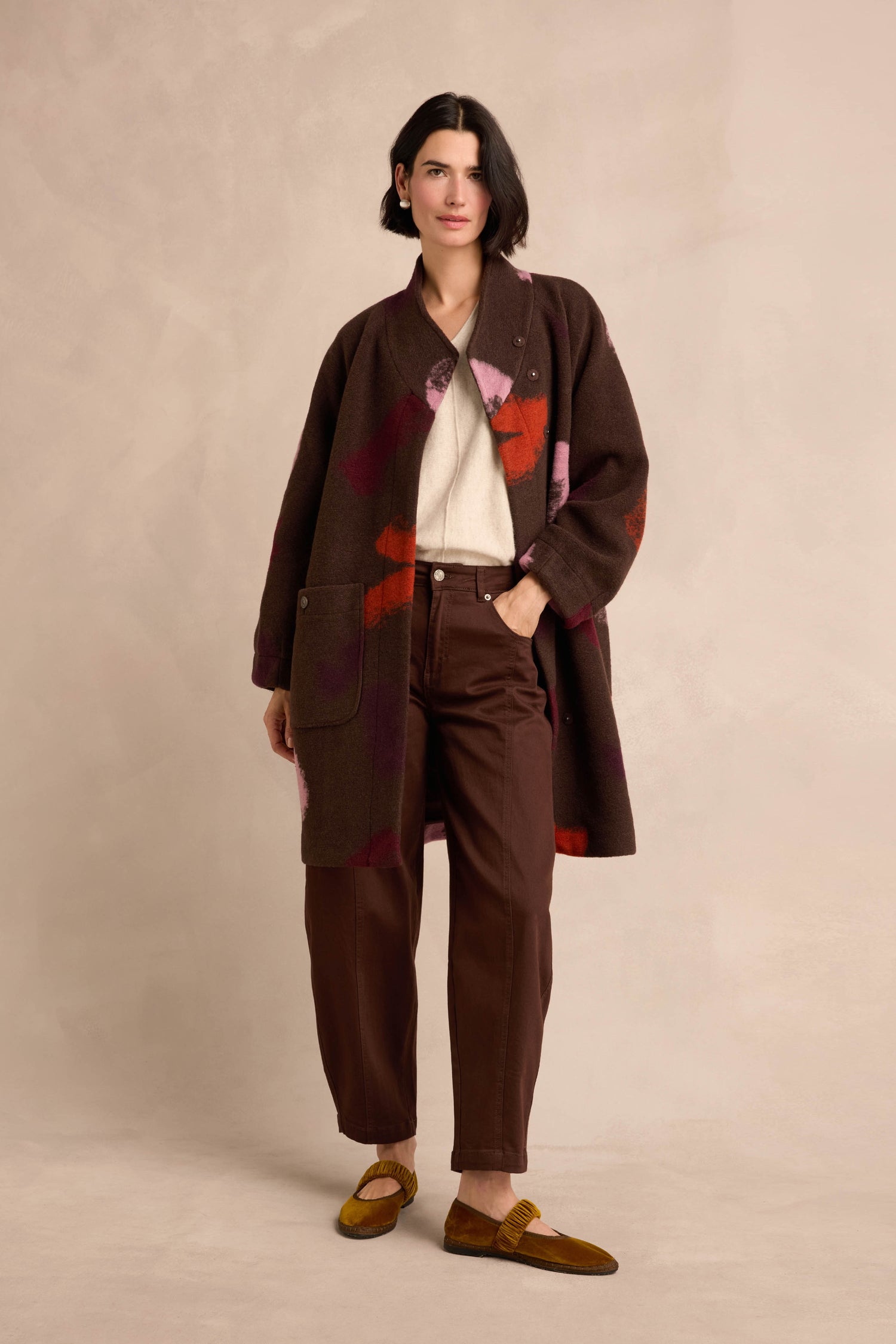 A person stands against a neutral background, wearing the Colour Splash Coat—a long winter coat with a patterned brown design—styled over a white top, brown pants, and yellow shoes.