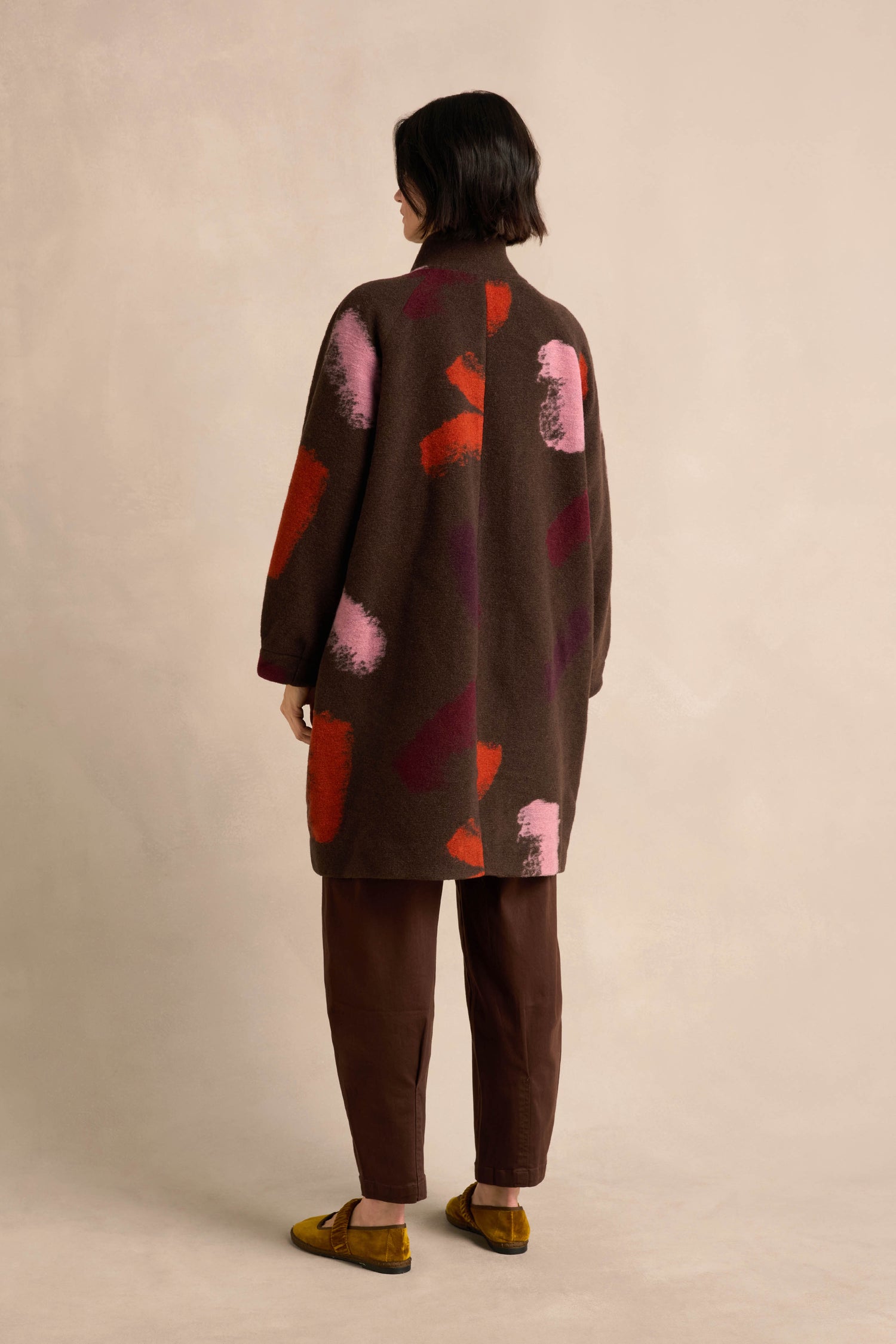 A person stands with their back to the camera, wearing the Colour Splash Coat—a long winter coat adorned with colorful abstract patterns—paired with brown pants and yellow shoes, set against a plain beige background.