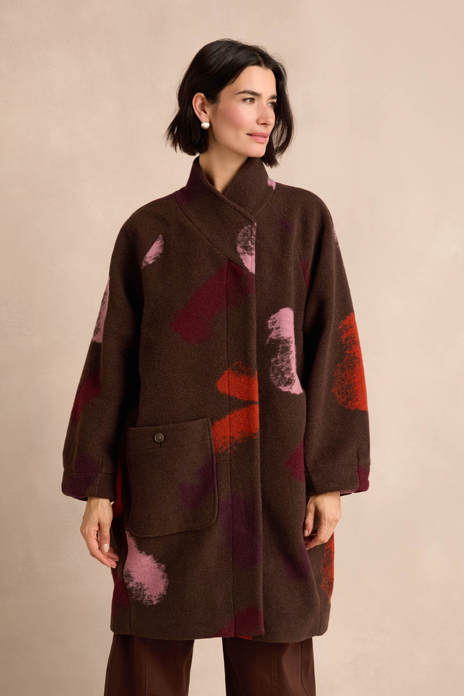 A woman stands against a plain beige background wearing the Colour Splash Coat—a loose, wool blend design featuring red and pink abstract patterns and a large front pocket—paired with dark pants.