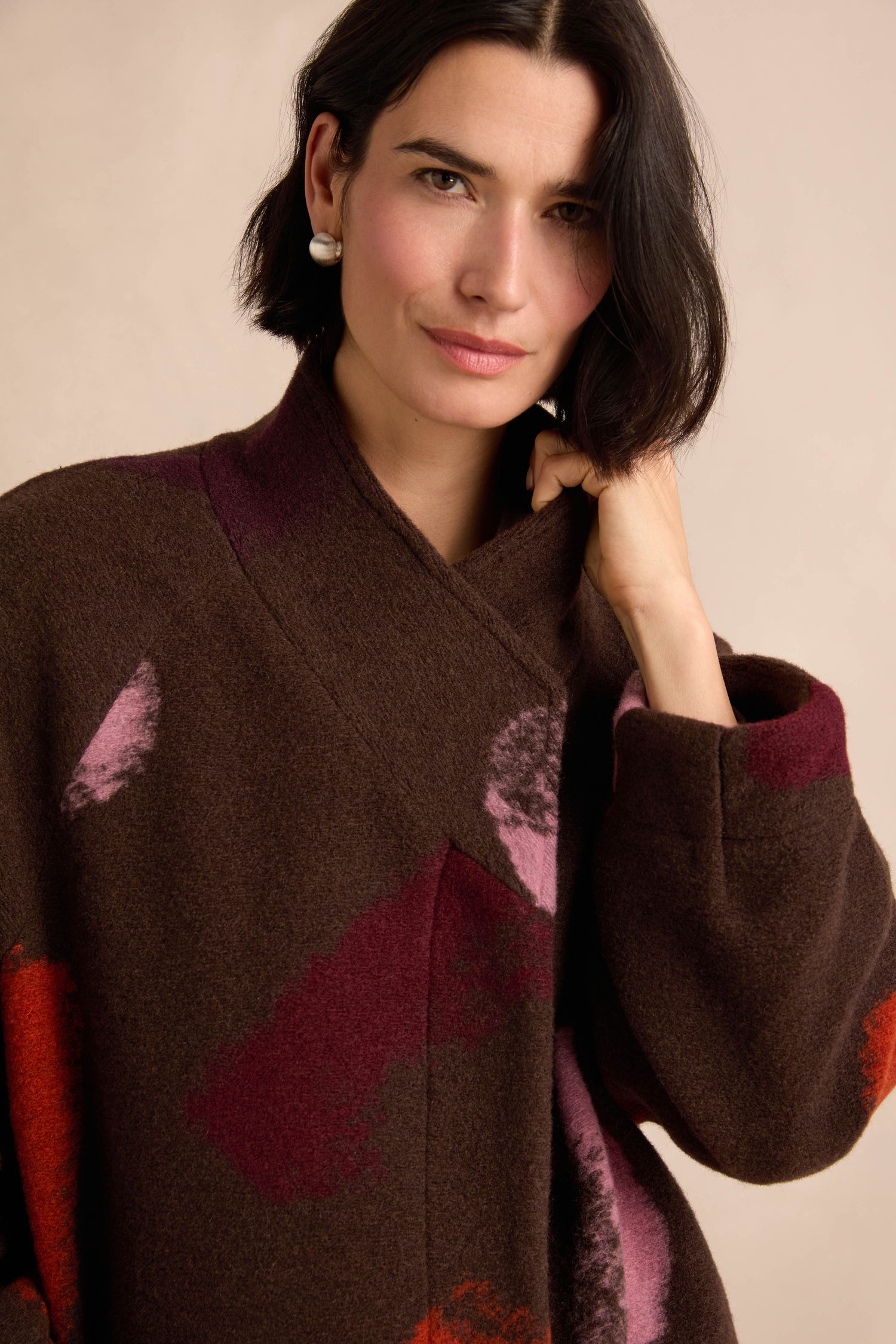 A woman with short dark hair wears the Colour Splash Coat—a long winter coat with a brown pattern highlighted by red and pink accents—looking at the camera with one hand touching her collar.