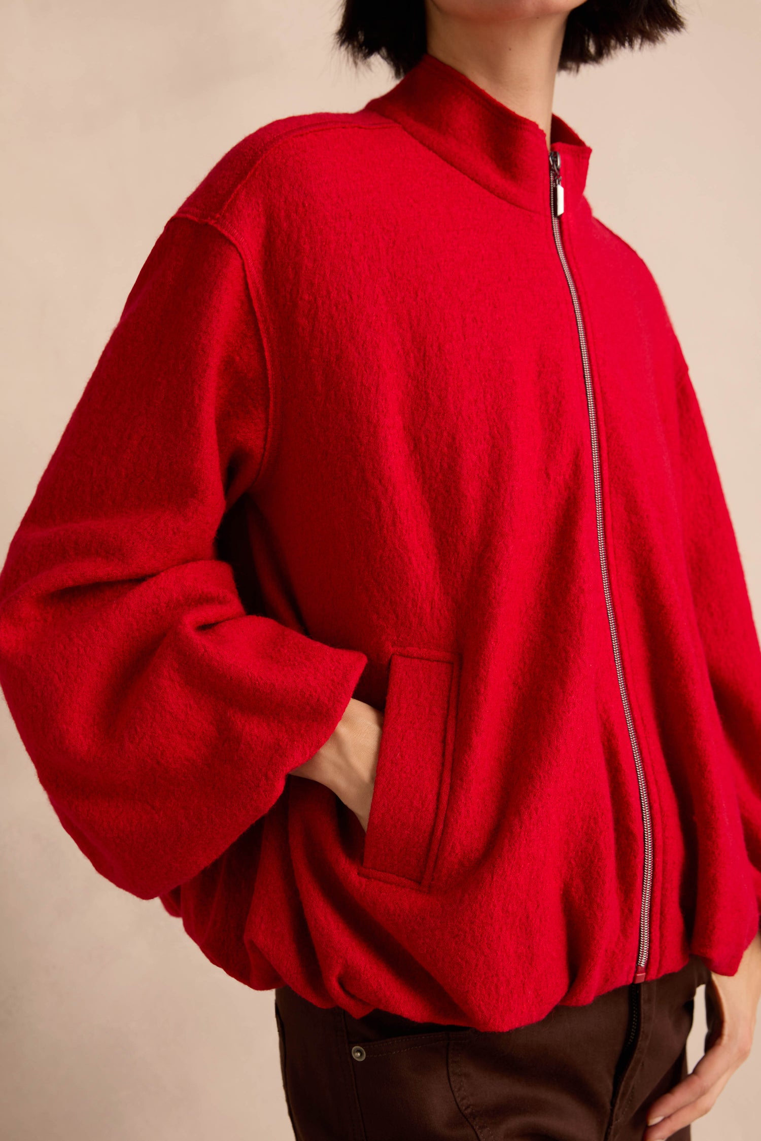 Wearing the Puffball Boiled Wool Jacket—a loose-fitting, bright red zip-up—hands tucked in the pockets, shown from shoulders to hips against a neutral background.