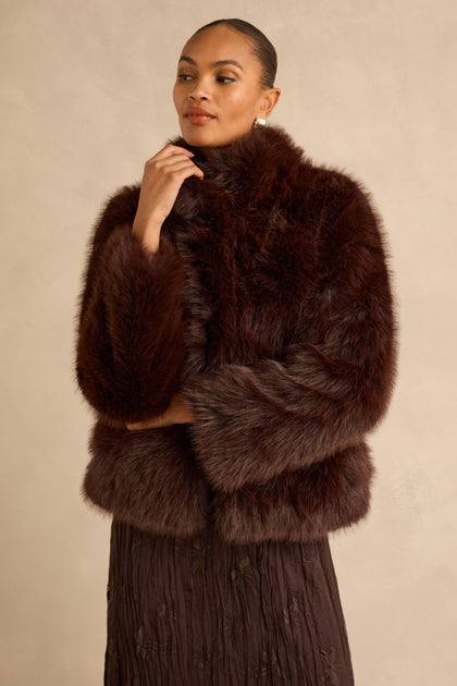 A person wearing a chocolate Faux Fur Jacket with a high stand collar over a textured brown skirt.