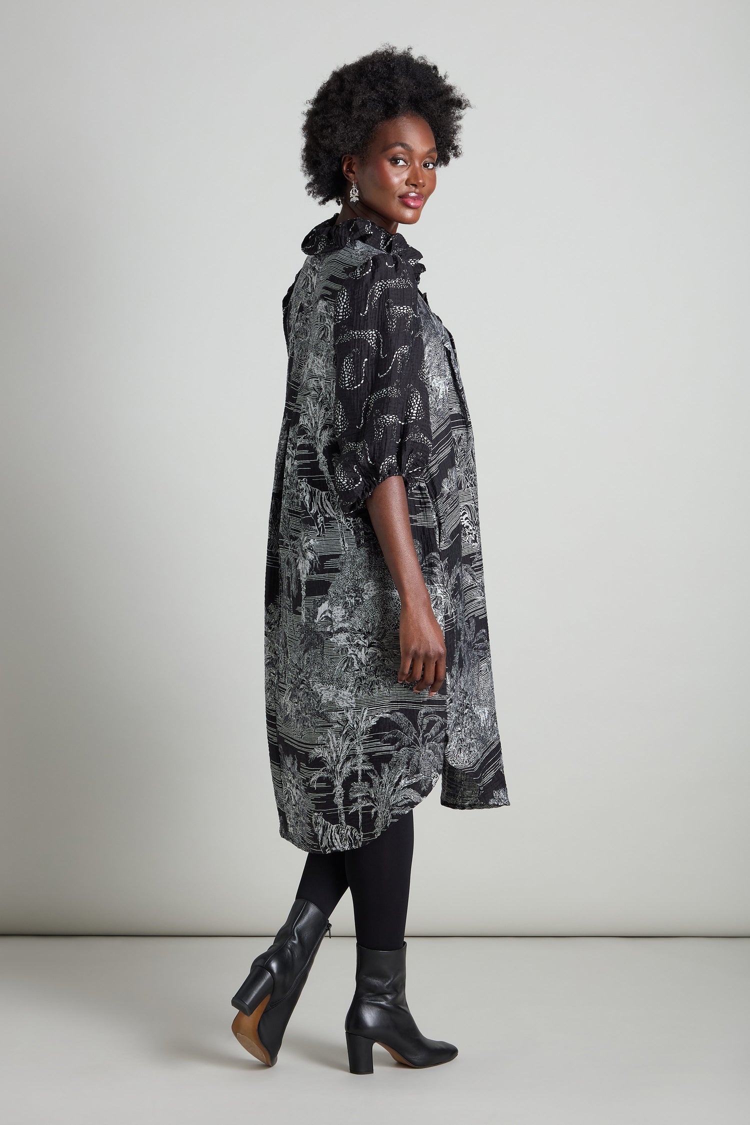 A person poses sideways, highlighting an A-line silhouette in a Frill Neck Viscose Dress with a black and white pattern, complemented by black leggings and heeled boots. The outfit subtly suggests a jungle-inspired print against a simple background.