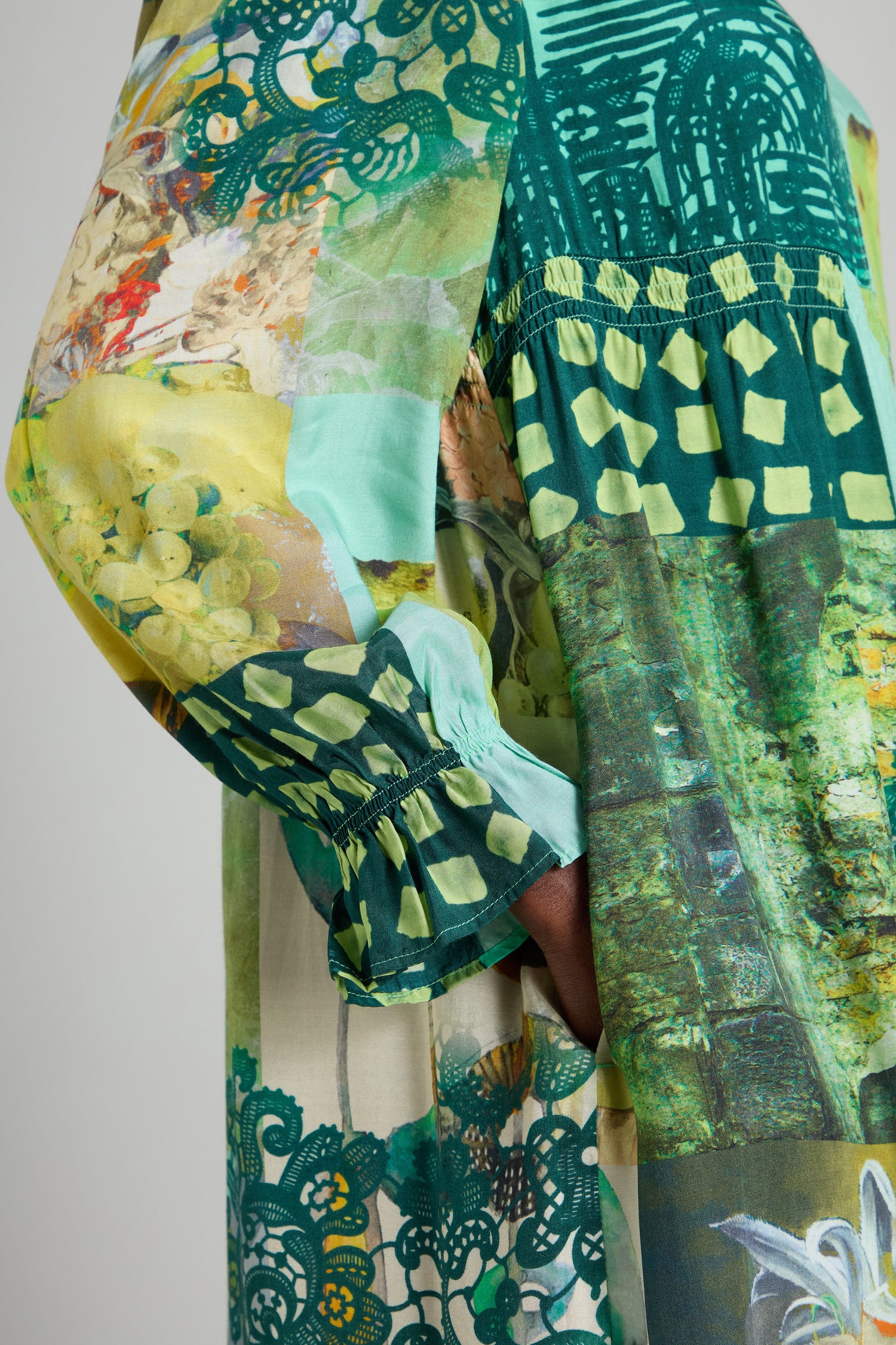 A close-up of someone wearing the Patchwork Print Viscose Dress, showcasing its vibrant patchwork designs in shades of green and yellow, with floral and geometric patterns. Their hand is tucked into the dress pocket.