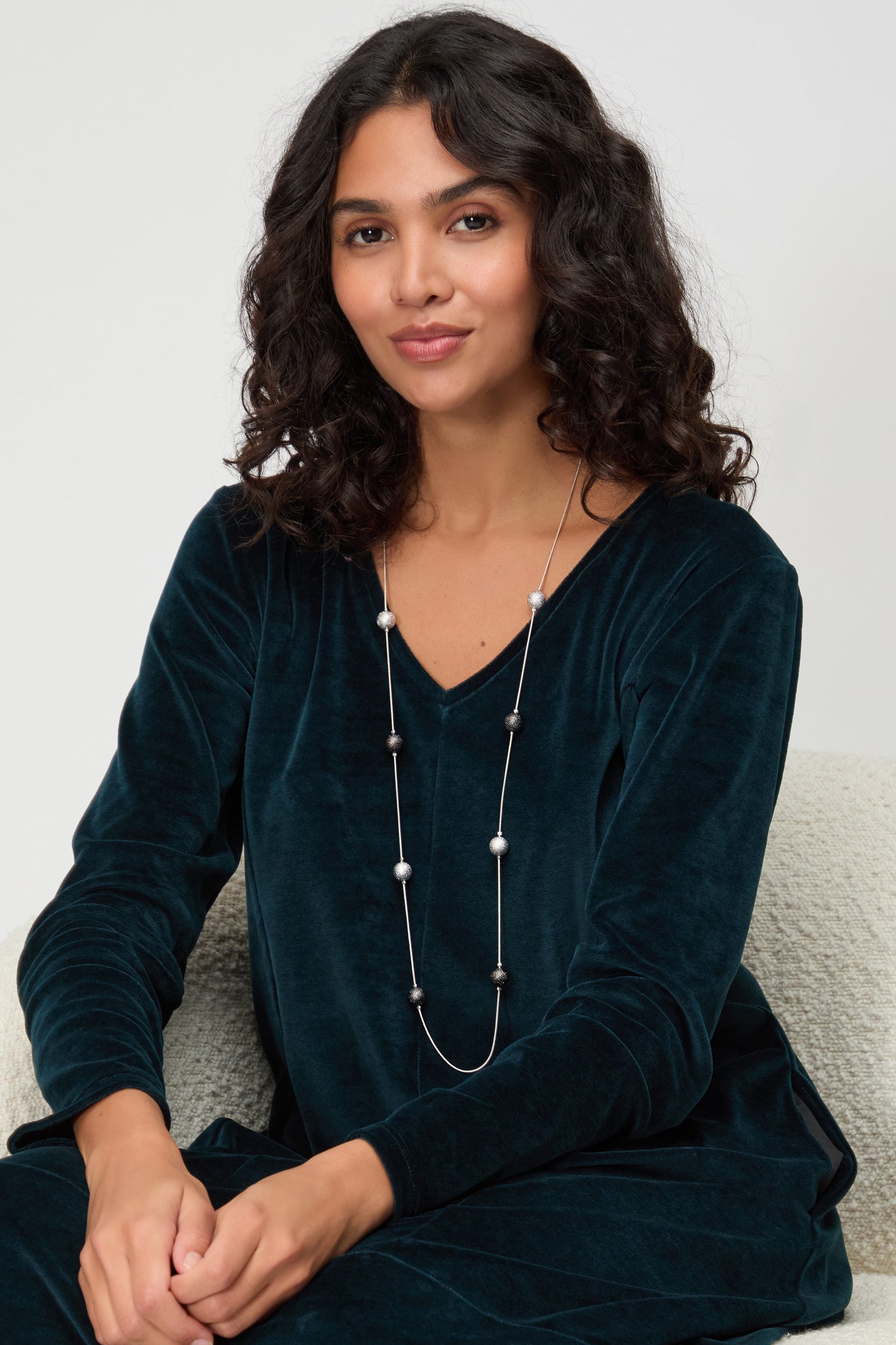 A woman with curly hair sits on a light chair, wearing a dark teal velvet top and the Sparkle Spheres Necklace, featuring silver and black beads, looking at the camera with a neutral expression.