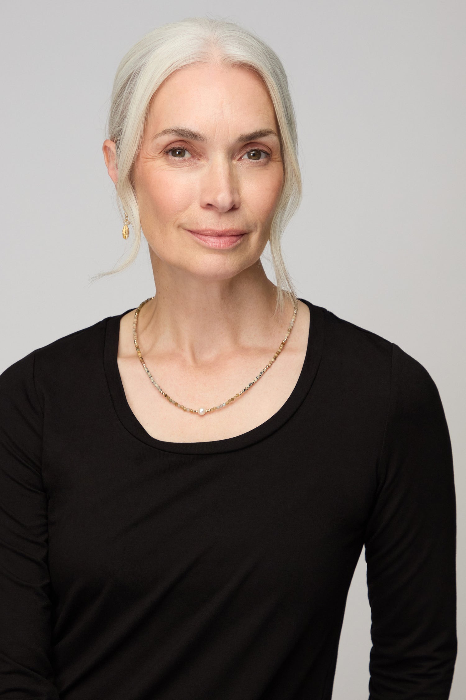 A woman with long gray hair in a low ponytail, wearing a black top, gold earring, and the Short Pearl Bead Necklace, faces the camera against a plain light background.