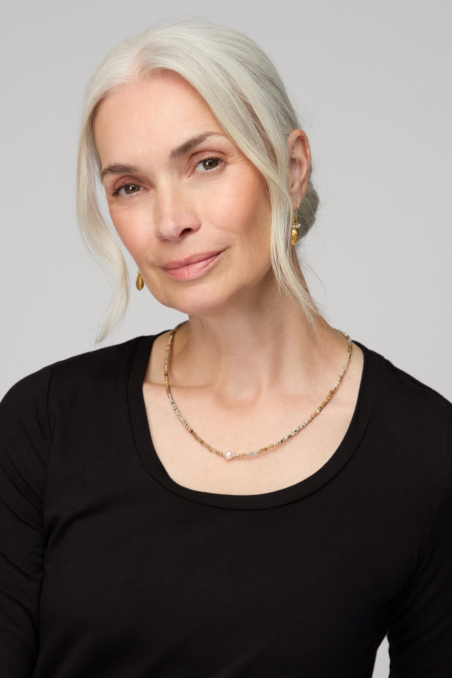 Woman with long gray hair wears a black top, gold earrings, and the Short Pearl Bead Necklace, looking at the camera against a plain background.