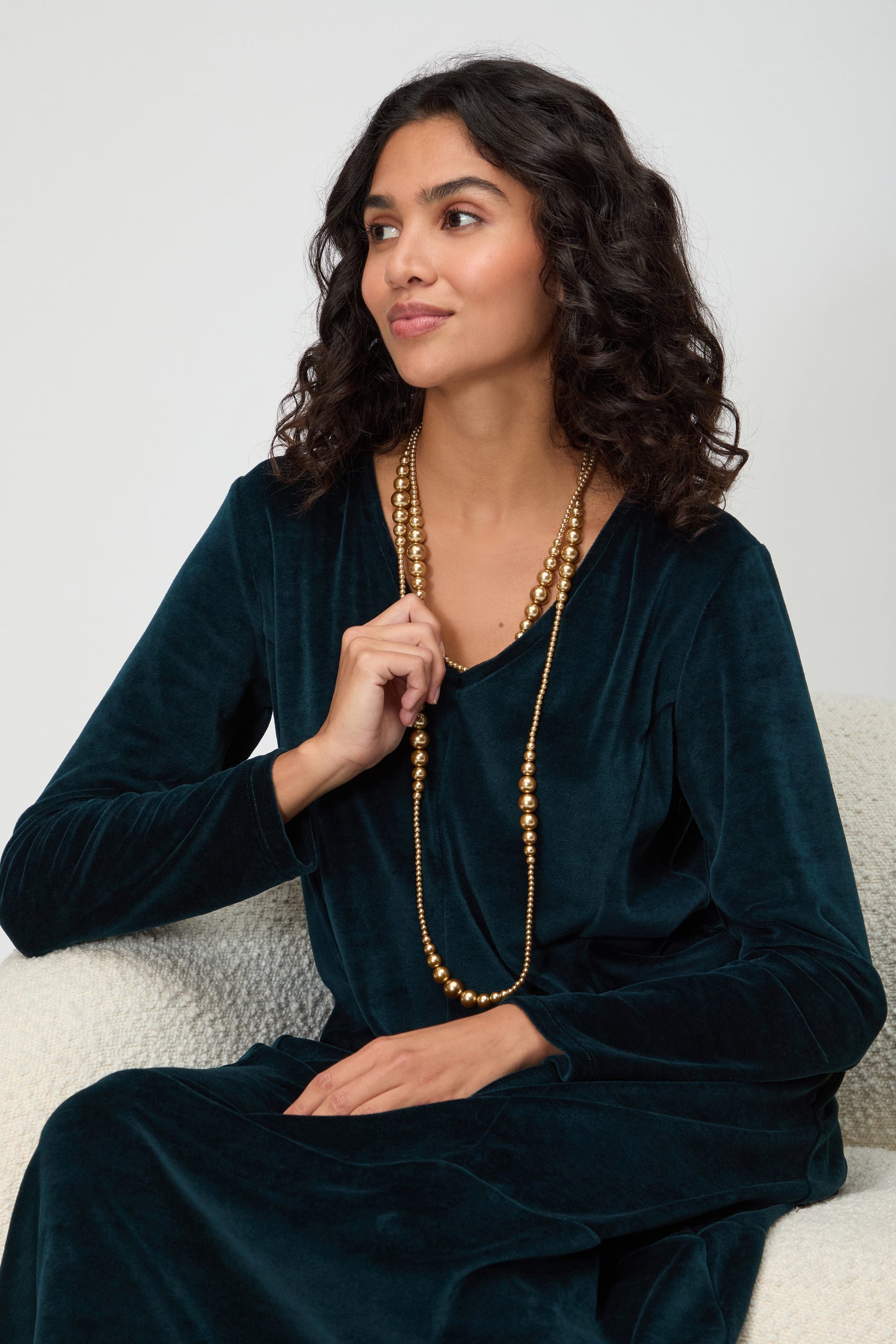 A woman with curly hair in a dark green velvet dress sits on a light chair, holding the Graduated Pearl Necklace and looking to the side.