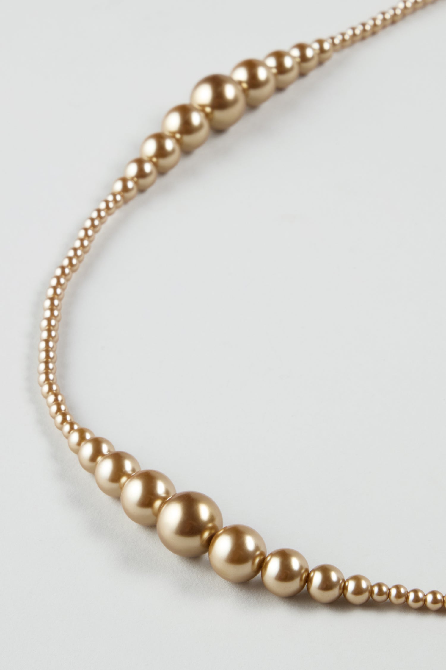 The Graduated Pearl Necklace showcases gold-toned beads in varying sizes, elegantly arranged in a modern pattern against a light background.