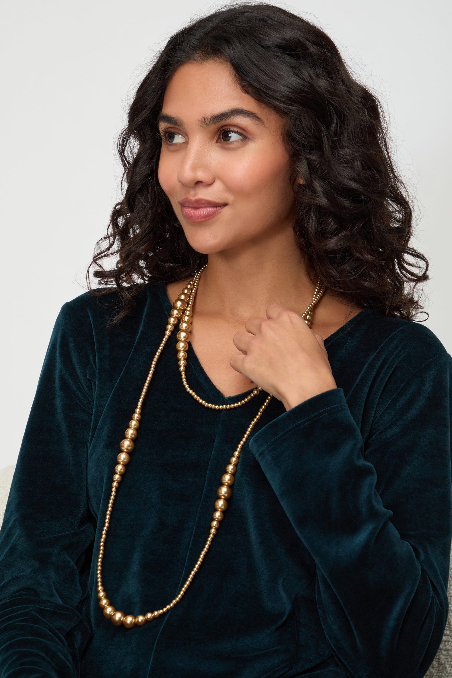 A woman with wavy dark hair wears a dark green velvet top and the Graduated Pearl Necklace, seated and gazing to the side.
