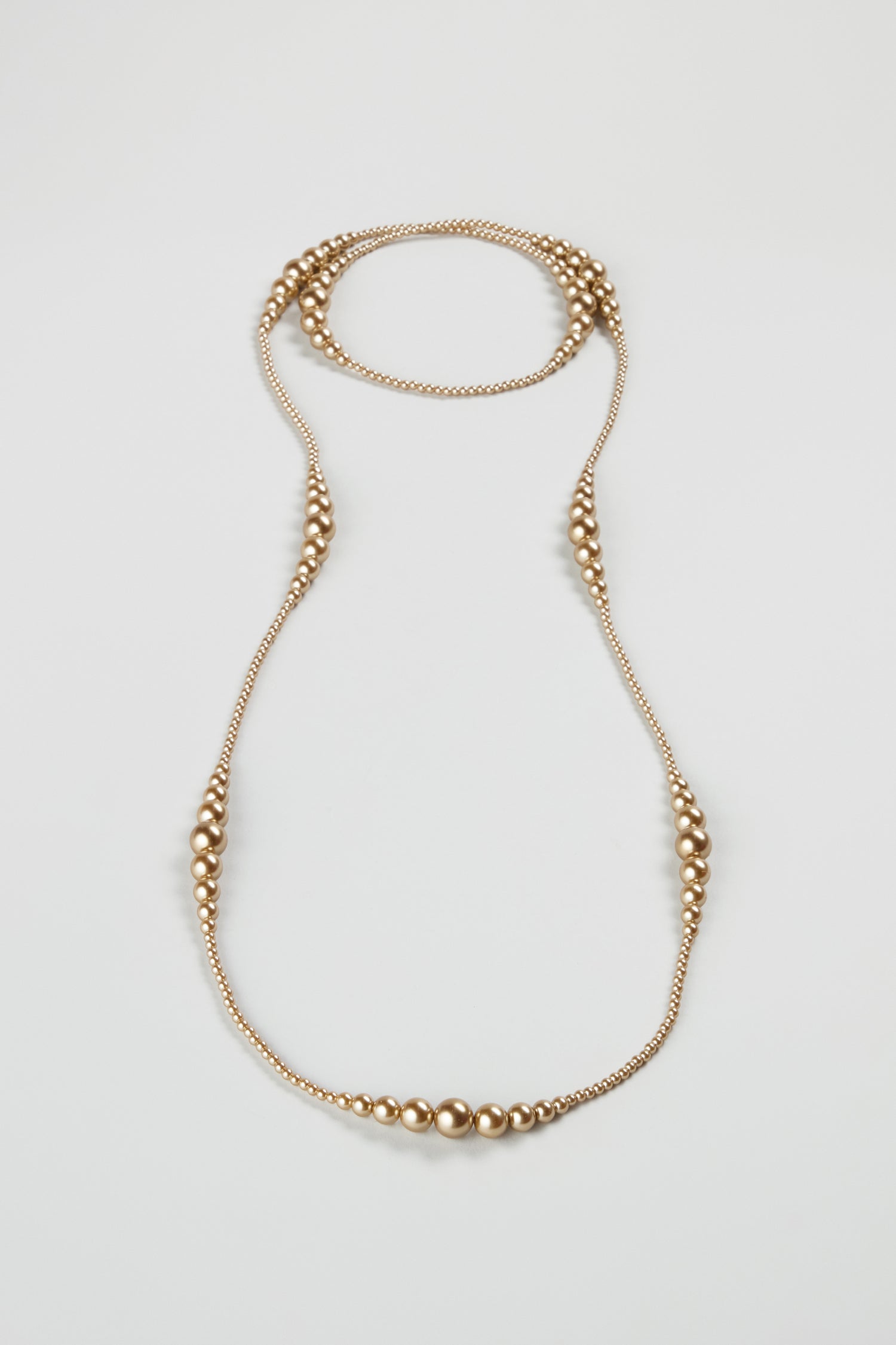 The Graduated Pearl Necklace features small and large round gold-colored beads in a symmetrical pattern, arranged as a long strand and displayed flat on a light background.