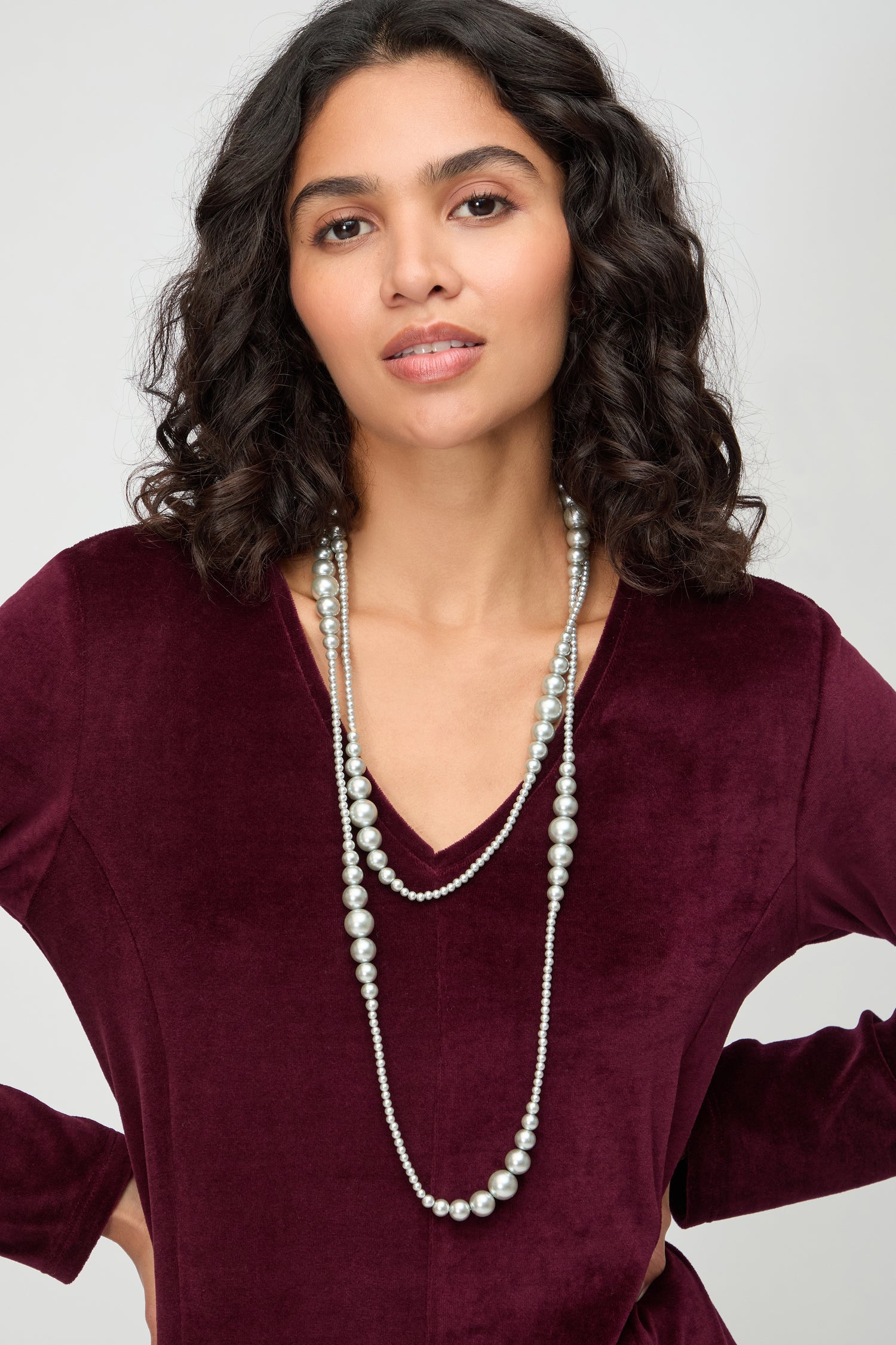 A woman with wavy dark hair wearing a burgundy velvet V-neck top and the Graduated Pearl Necklace—a long, double-strand modern pearl piece—stands against a plain light background.