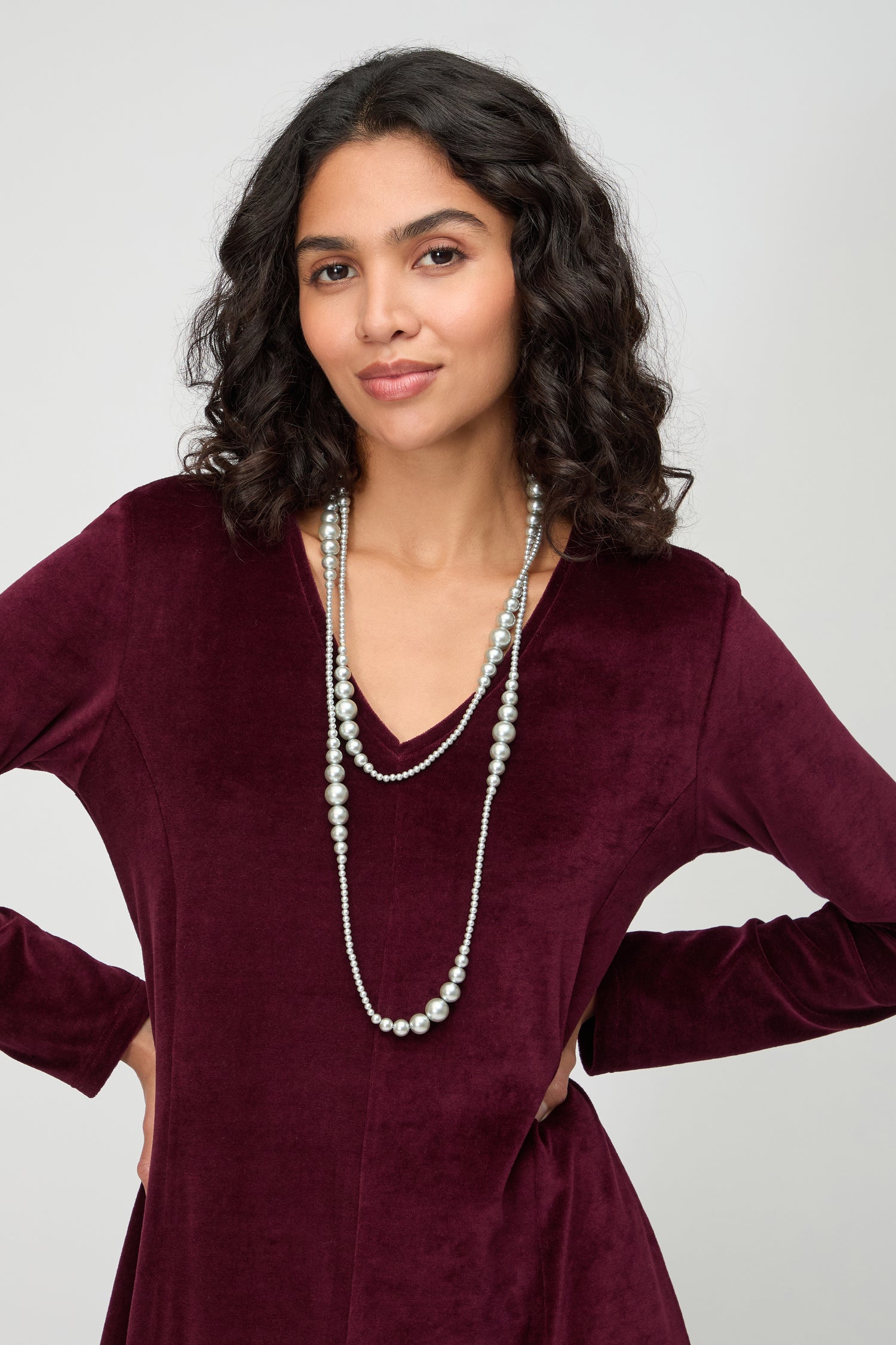 A woman with curly hair and hands on hips showcases the Graduated Pearl Necklace, styled with a burgundy velvet dress, against a plain background.
