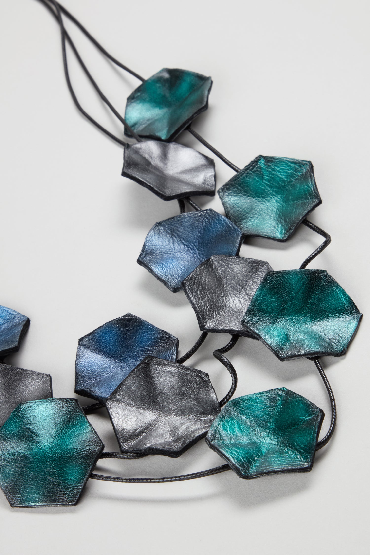 The Metallic Leather Shapes Necklace showcases handcrafted hexagonal leather pieces in green, blue, and silver tones, linked by black cords for a bold statement jewellery look—displayed against a light background.