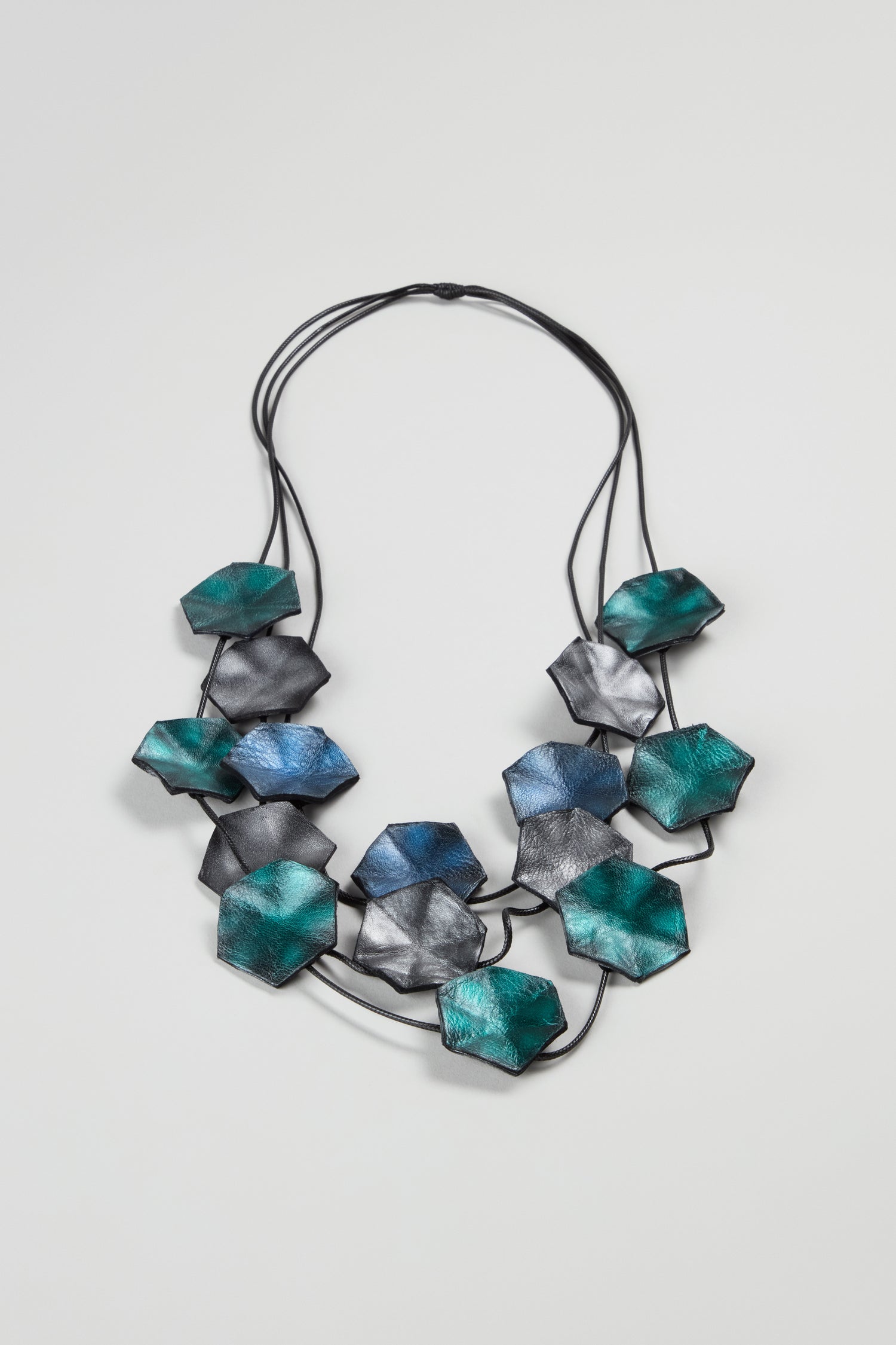 The Metallic Leather Shapes Necklace features three black cords with clustered geometric metallic blue-green and silver beads—a striking handcrafted statement piece for any occasion.