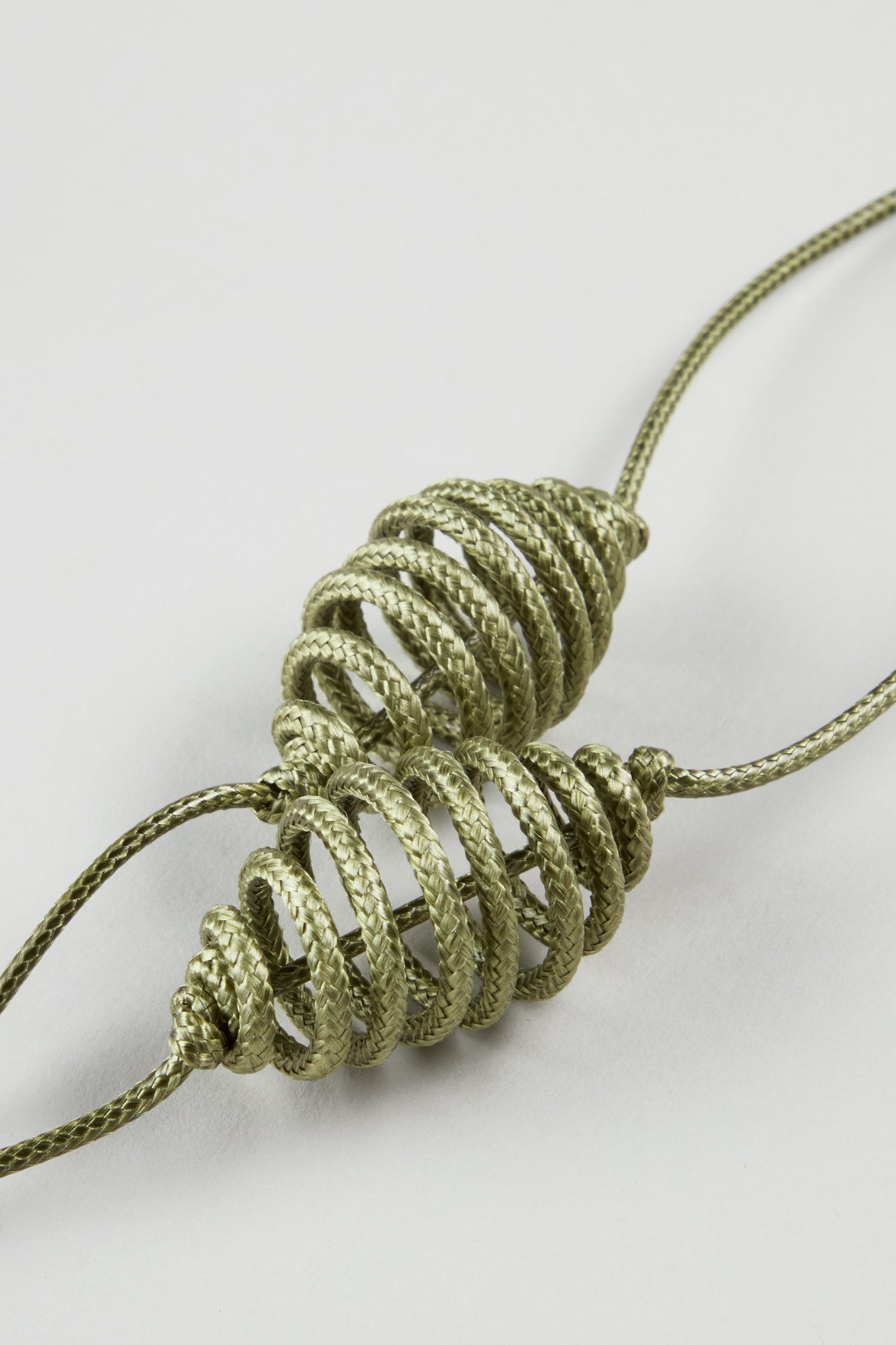 Two metallic cords of the Spiral Cord Khaki Necklace are artistically looped into spiral, cone-like shapes and placed parallel to each other on a plain white background.