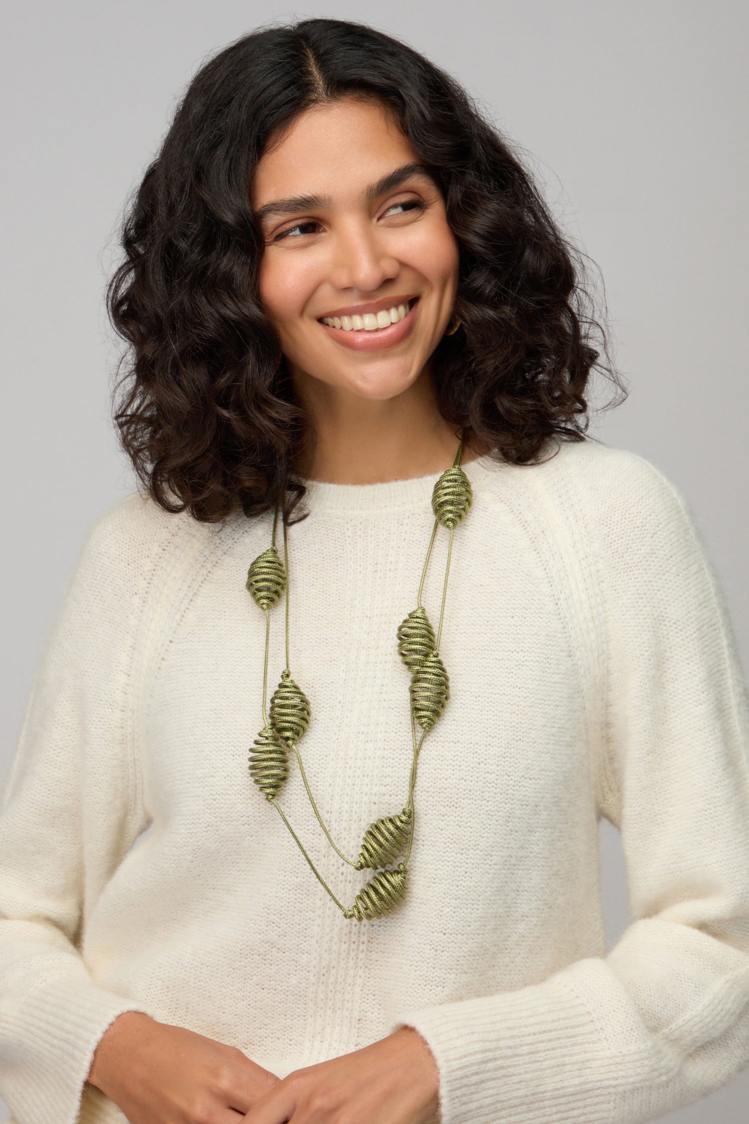 A woman with curly dark hair wears a white sweater and the Spiral Cord Khaki Necklace, smiling as she looks slightly to the side against a neutral background.