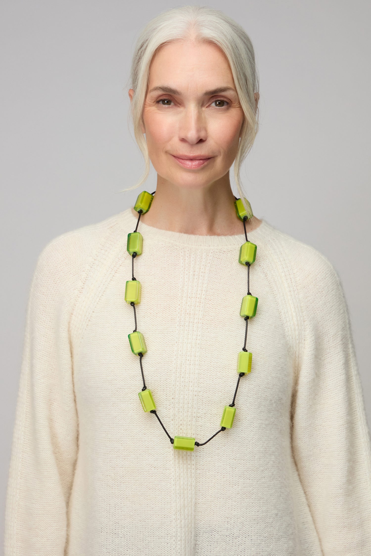 A woman with long gray hair in a cream sweater wears the Neon Resin Cubes Necklace, featuring large rectangular green resin cubes, and stands against a plain background.
