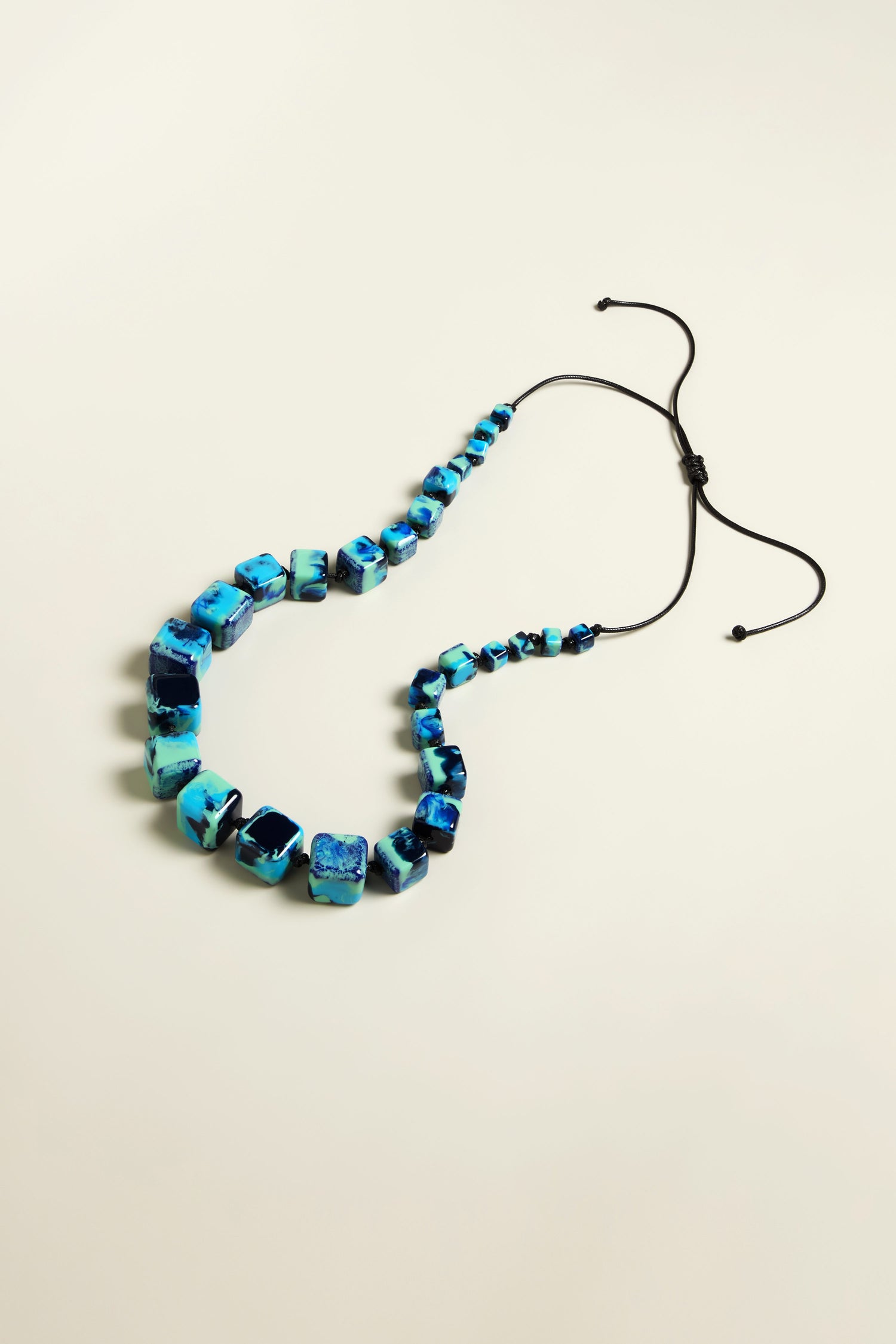 The Watercolour Cubes Necklace highlights blue and turquoise marbled square beads on an adjustable cord, with bead sizes increasing gradually from back to front.