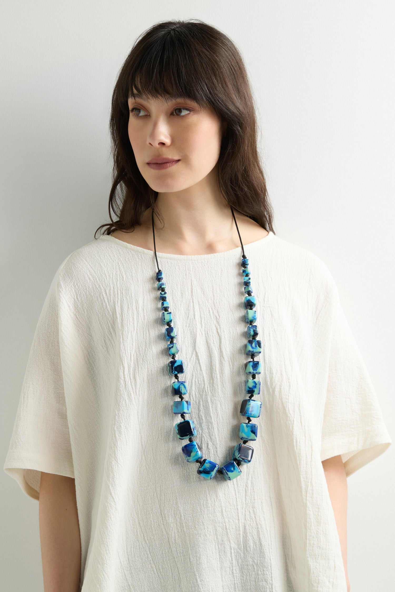 A woman with long dark hair in a white blouse wears the Watercolour Cubes Necklace, featuring an adjustable cord, against a plain white background.