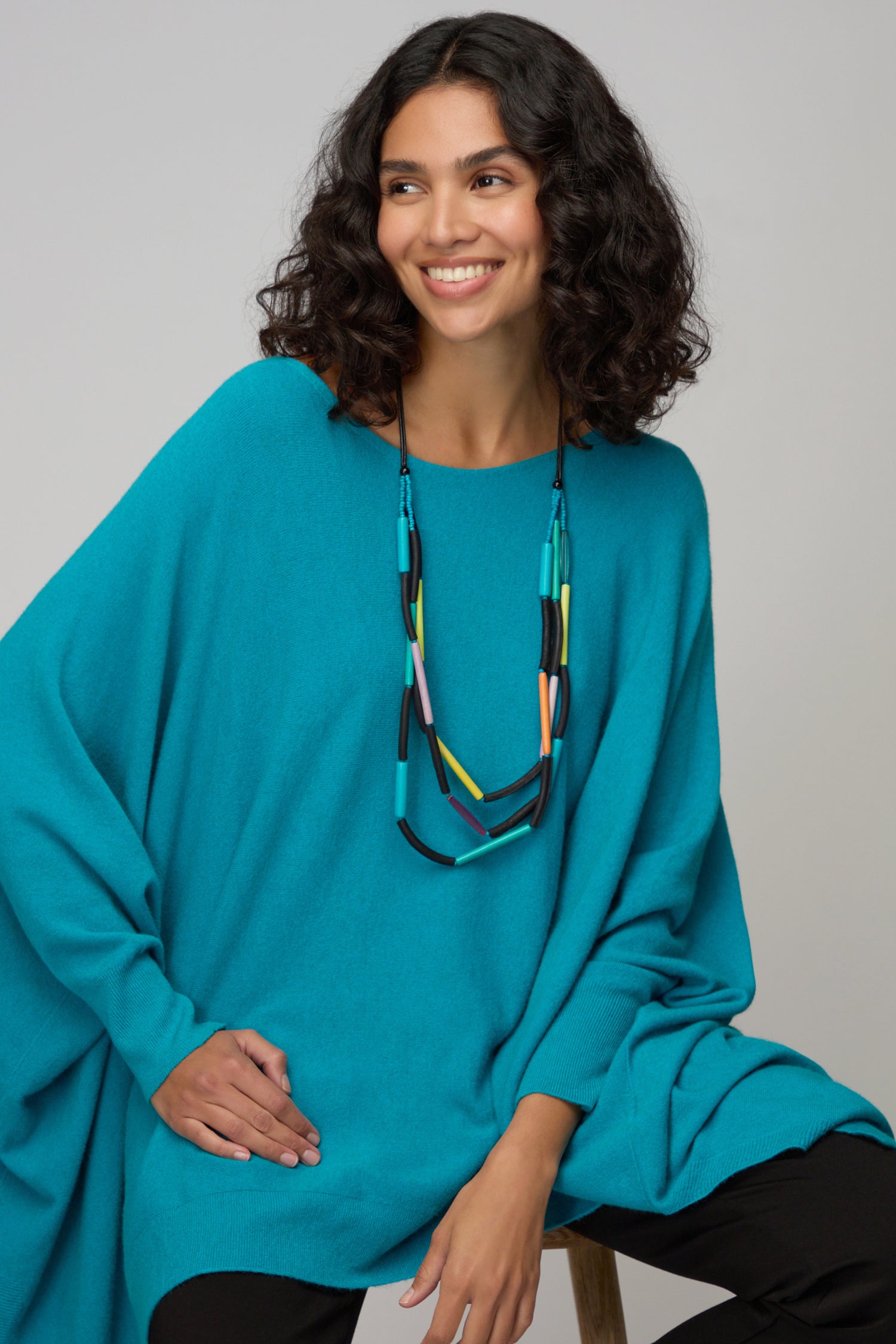 Woman with curly hair sits smiling, one hand on her knee, wearing a bright blue poncho and the Colourful Cylinders Necklace.