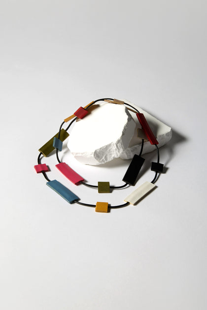 The Mondrian Necklace, showcasing a Bauhaus-inspired design, features colorful rectangular beads in red, blue, green, and white on a black cord, set against a plain gray background with a white rock backdrop.