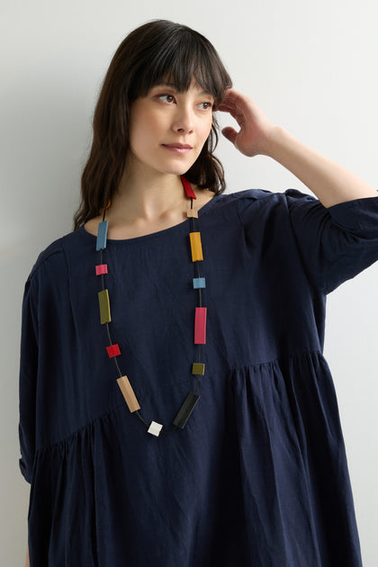 A person with long dark hair, wearing a navy dress and a Mondrian Necklace with colorful geometric shapes, stands against a plain white background looking to the side.