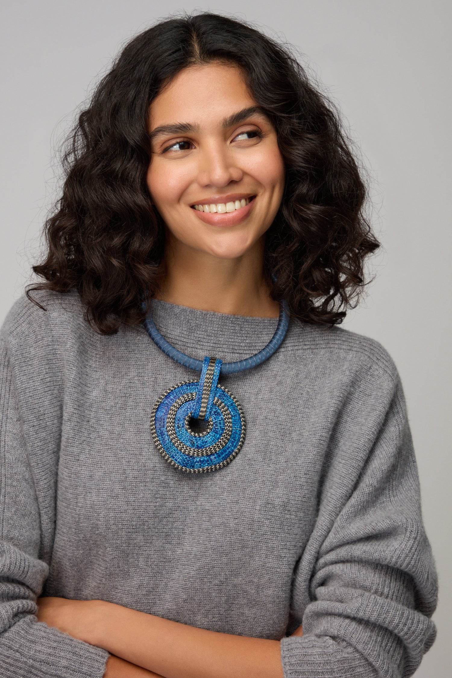 Woman with curly dark hair, wearing a grey sweater and the Woven Circle Necklace, smiles and looks slightly to the side against a plain background.