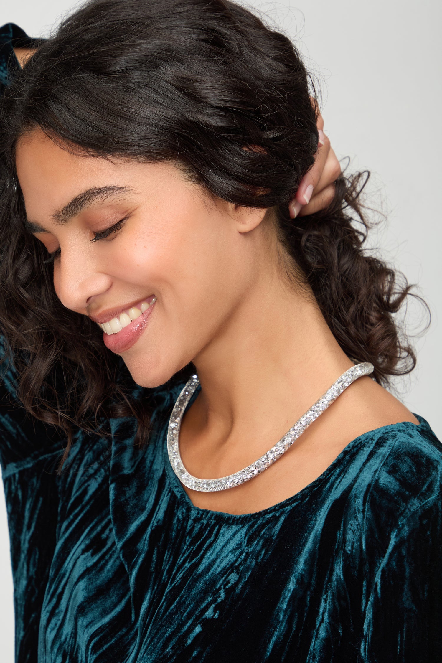 Woman with wavy dark hair, eyes closed and smiling with one hand behind her head, showcases the Short Silver Diamante Necklace while wearing a blue velvet top—capturing the allure of contemporary jewellery.