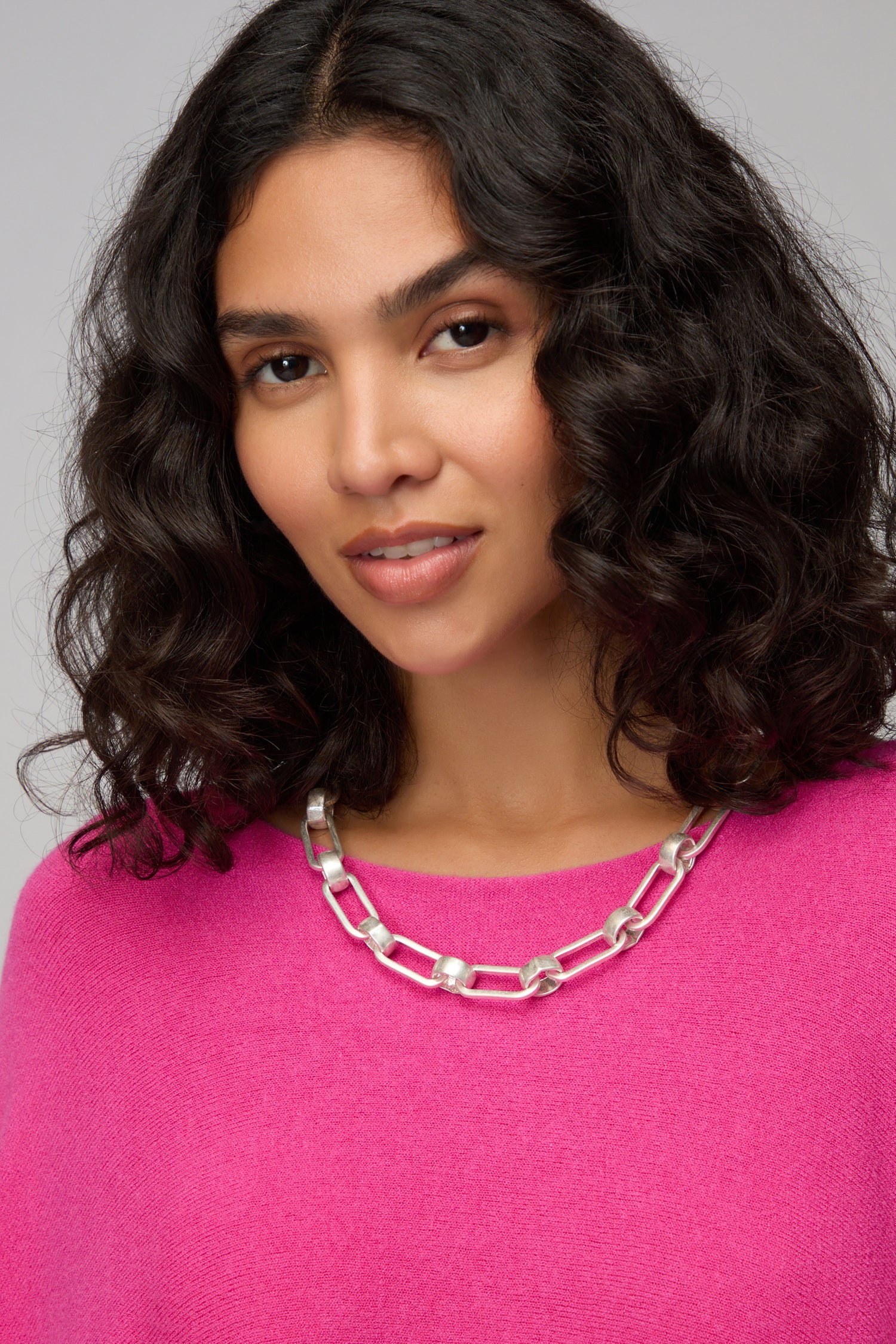 Woman with wavy dark hair wears a bright pink sweater and the Short Silver Links Necklace, looking at the camera with a neutral expression.
