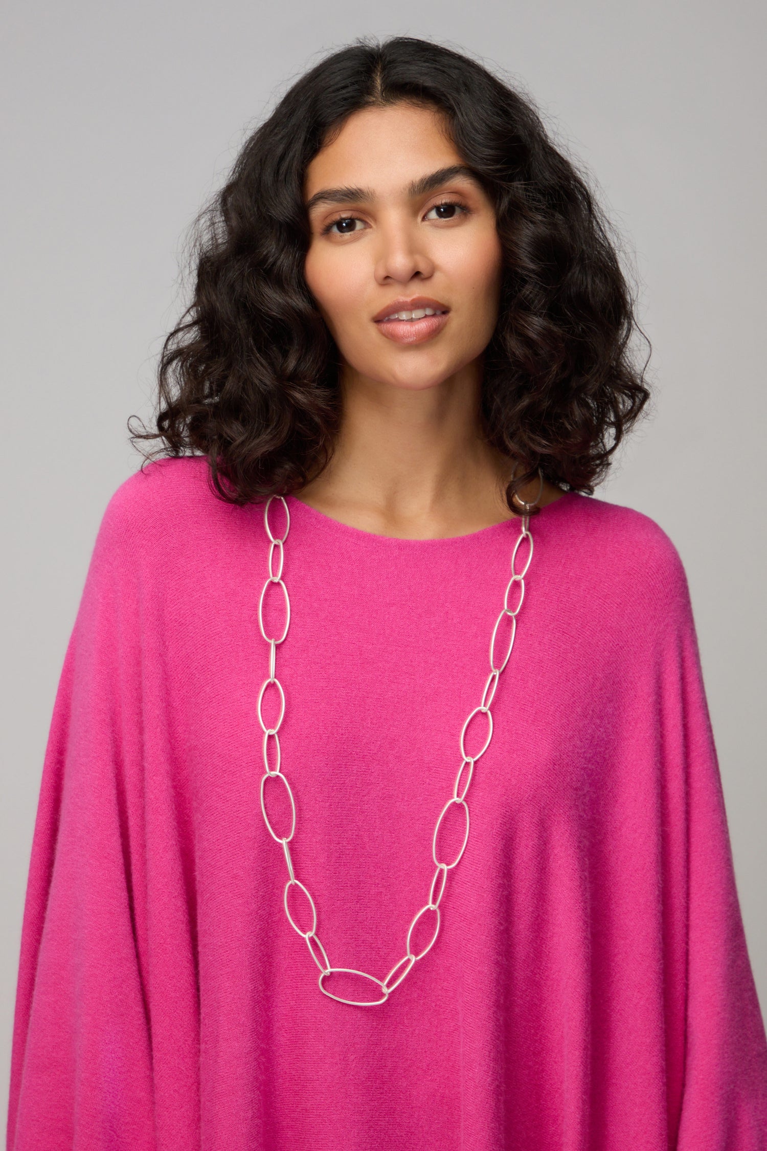 A woman with curly dark hair wears a bright pink top and the Long Silver Hoops Necklace, standing against a neutral background.