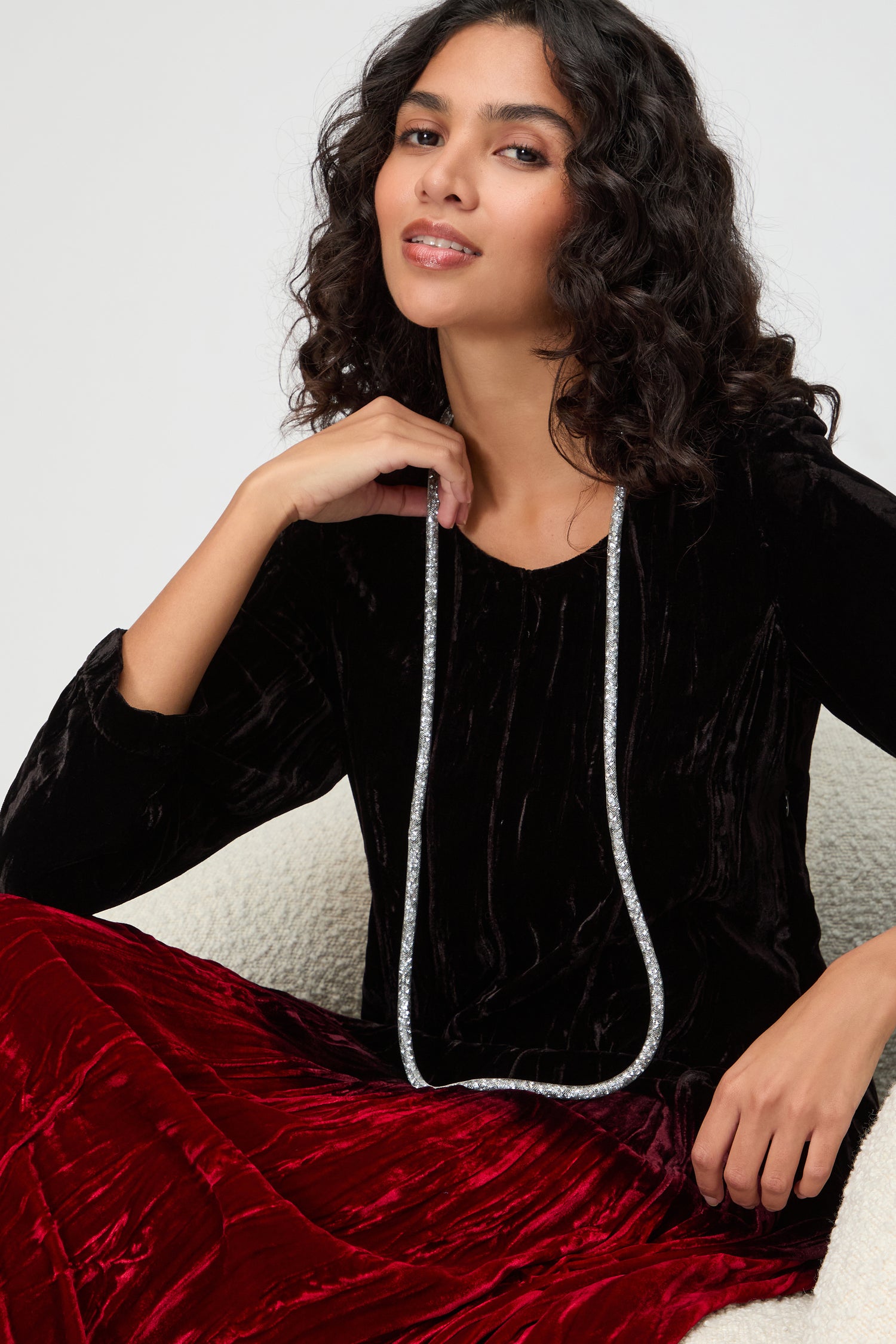 A woman with wavy dark hair sits on a chair, wearing a black velvet top, red velvet skirt, and a Long Silver Diamante Necklace that adds a statement touch to her elegant look.