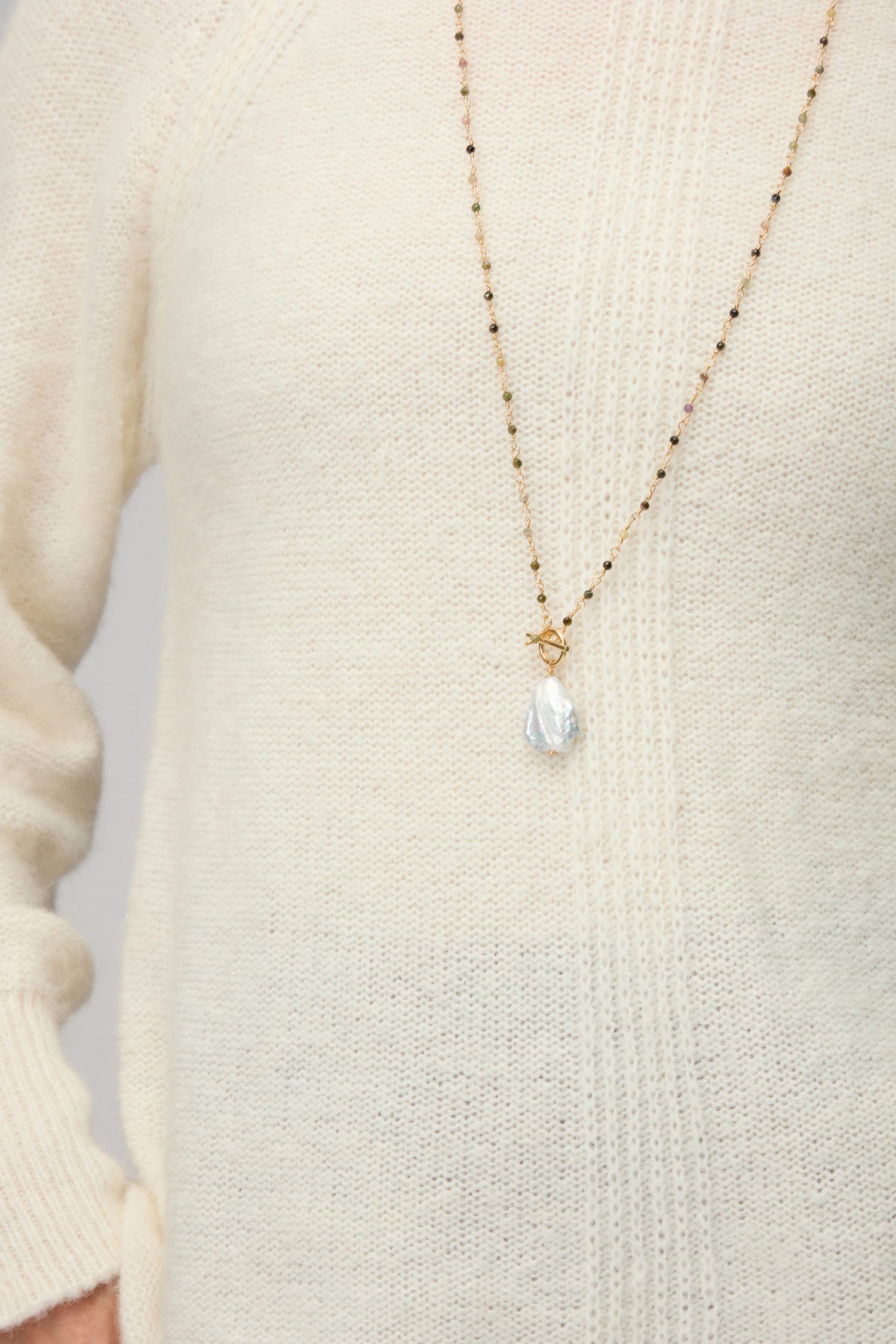 A person wearing a cream knit sweater paired with the Long Freshwater Pearl Necklace featuring a gold chain and a teardrop-shaped pearl pendant.