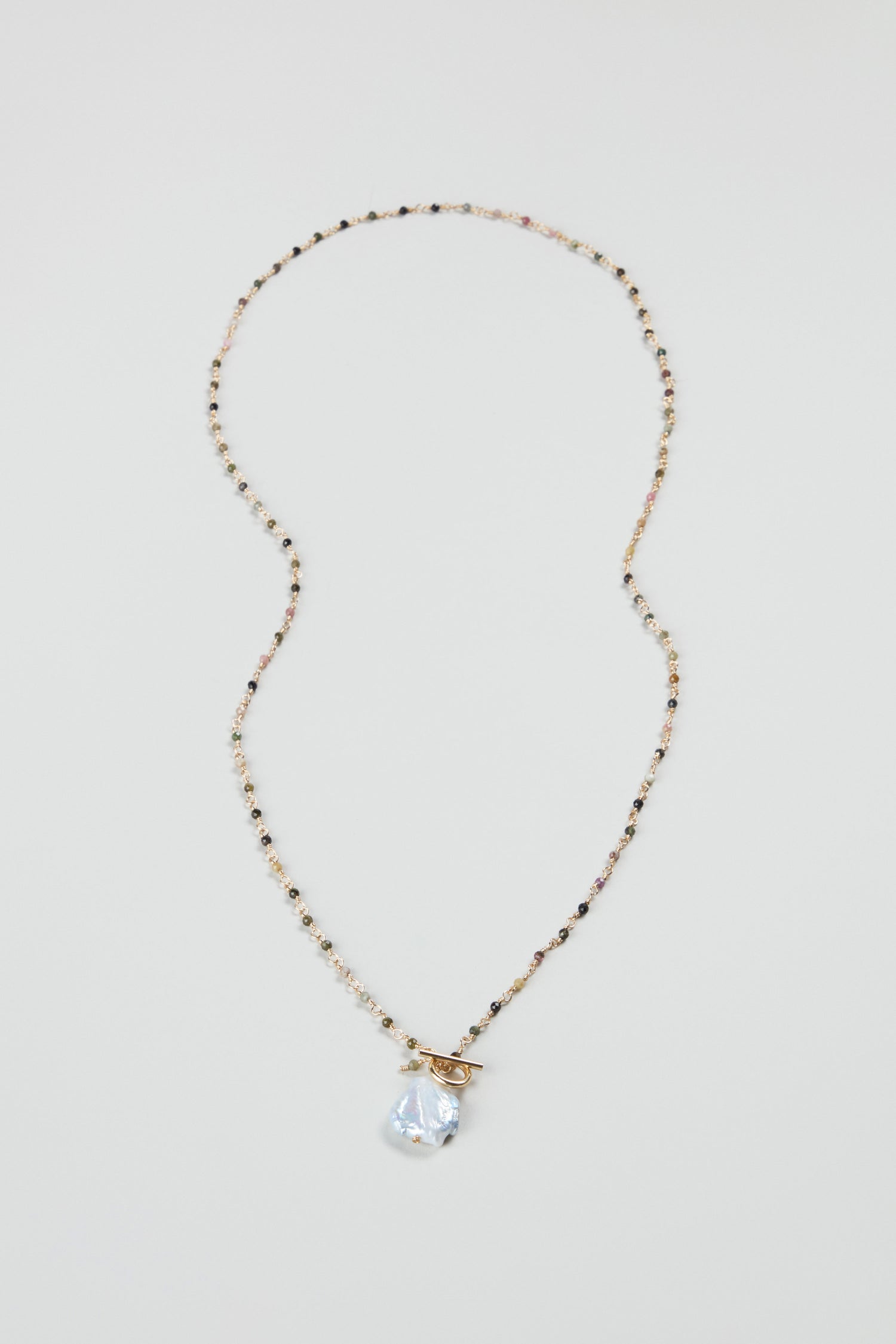 The Long Freshwater Pearl Necklace features delicate beads, a clear teardrop pendant, and a gold toggle clasp, beautifully displayed on a plain light background.