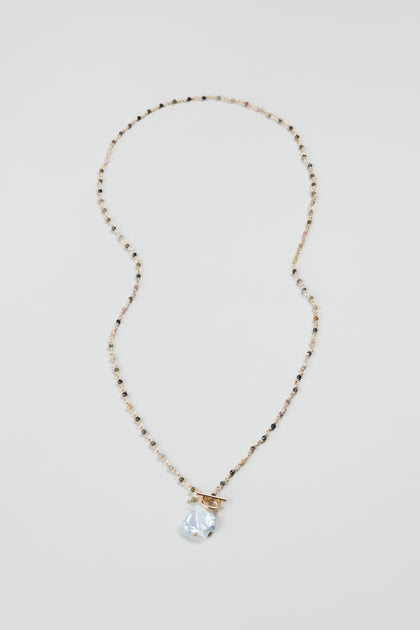 The Long Freshwater Pearl Necklace features delicate beads, a clear teardrop pendant, and a gold toggle clasp, beautifully displayed on a plain light background.