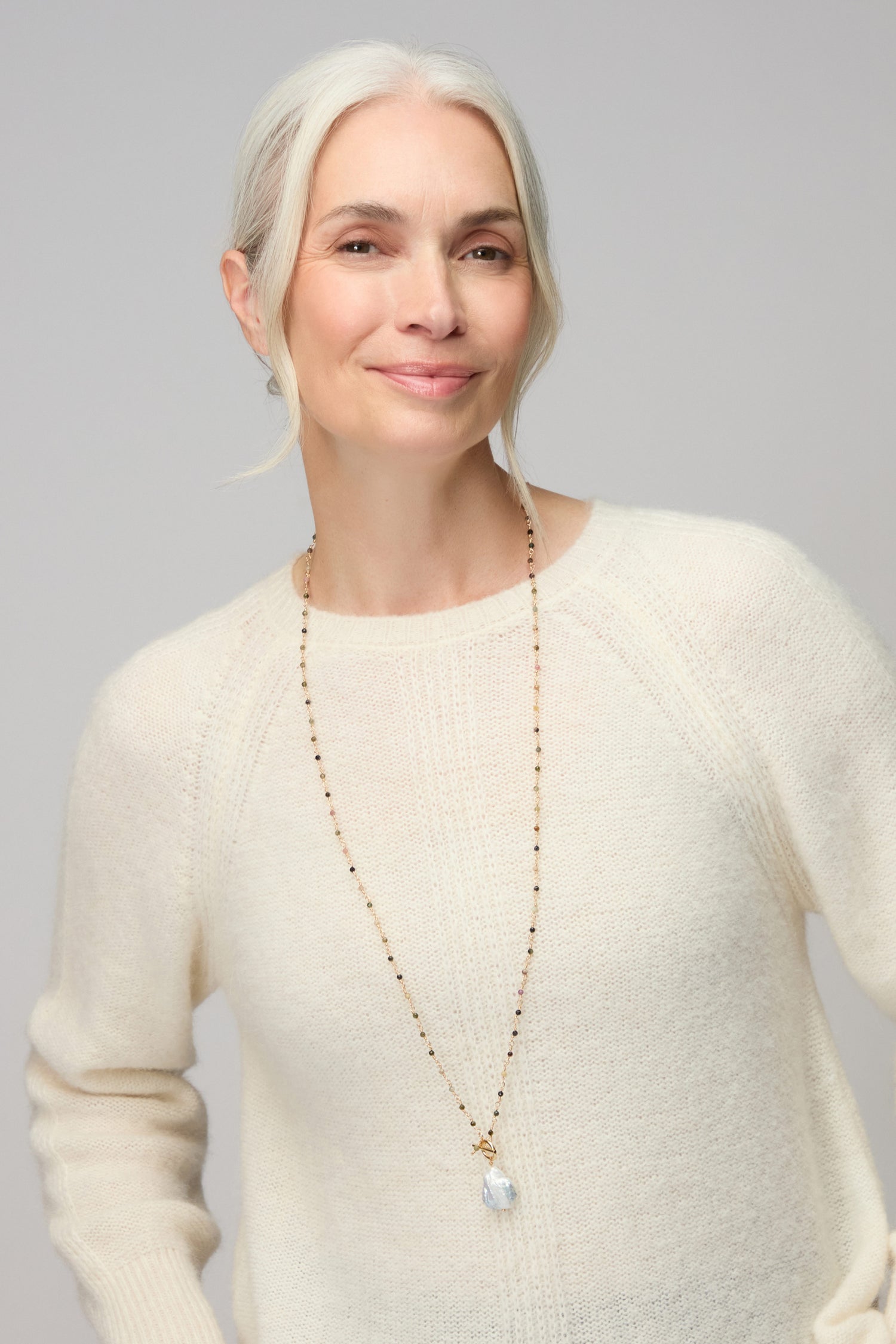 A woman with gray hair in a low bun models the Long Freshwater Pearl Necklace, paired with a cream sweater, against a simple light background.