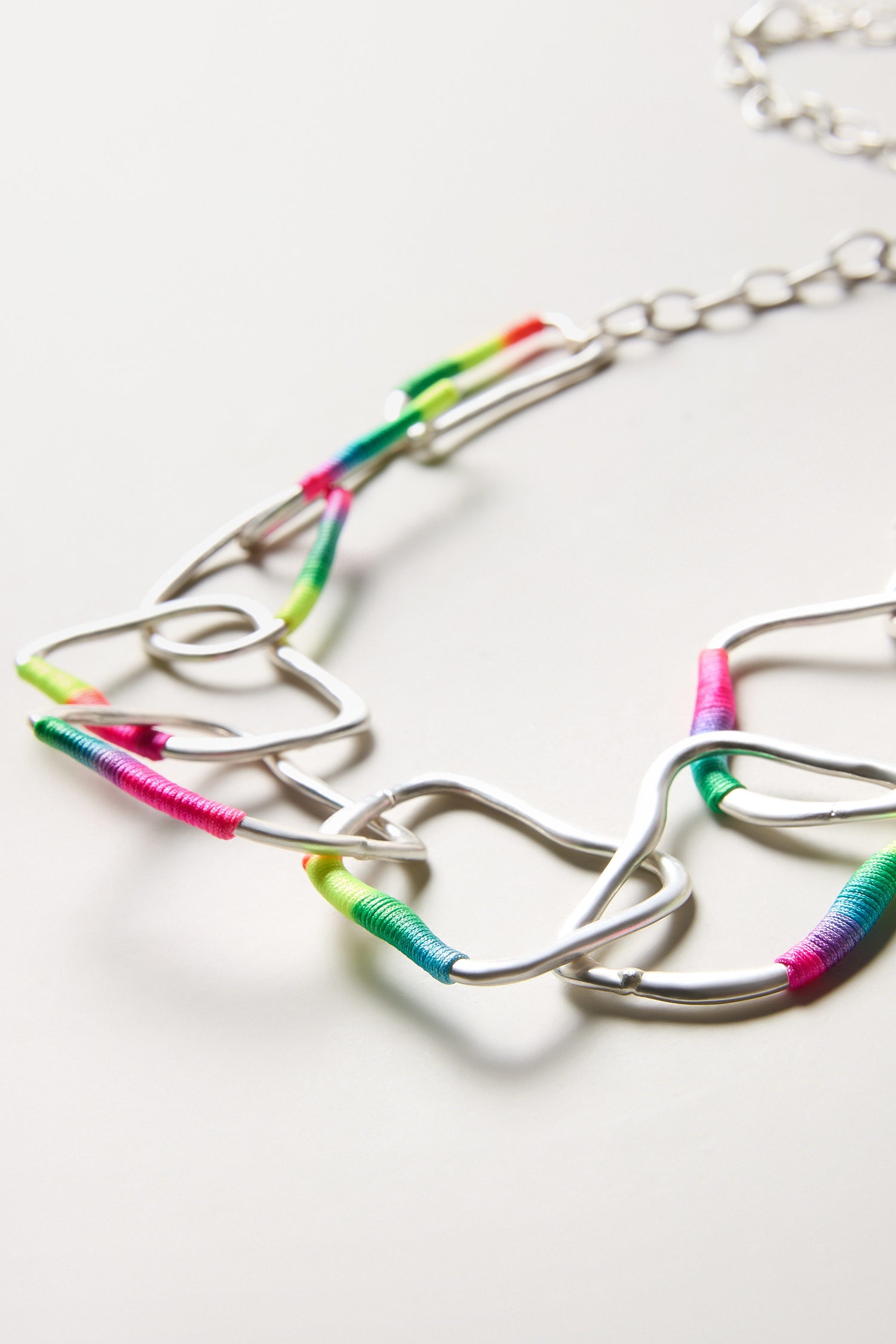 The Rainbow Detail Crushed Hoop Necklace features irregular silver links with a crushed finish, partially wrapped in multicolored thread for a vibrant statement look, shown on a white surface.