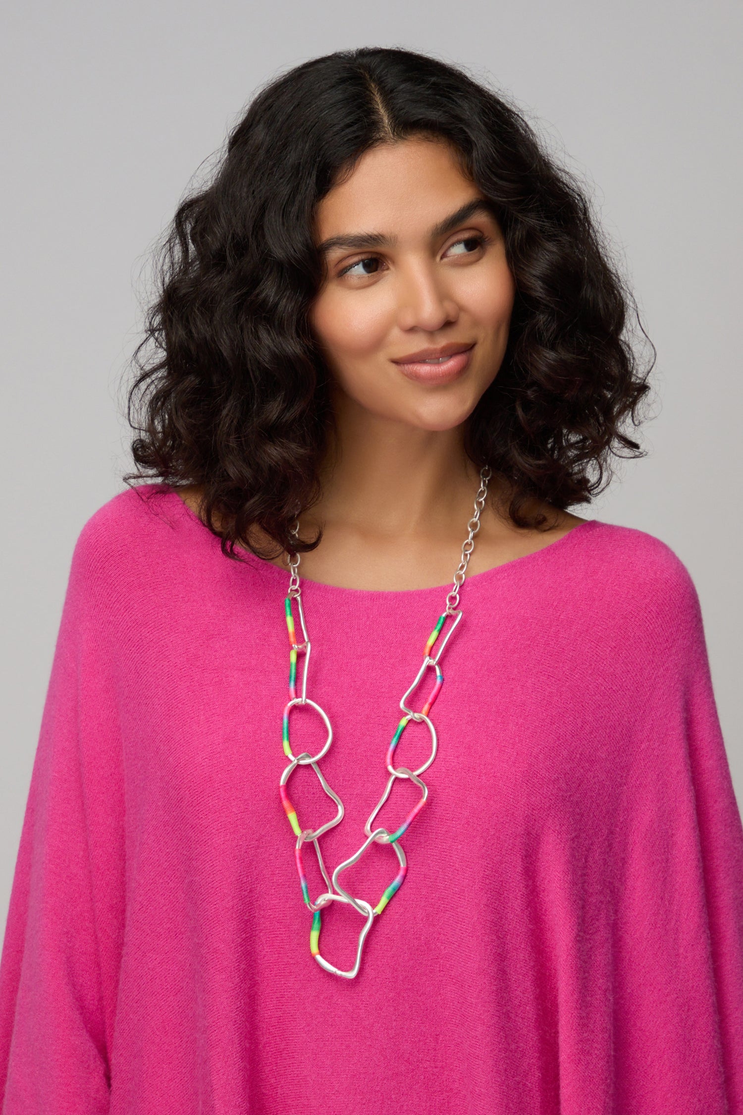 Woman with curly dark hair wears a bright pink top and the Rainbow Detail Crushed Hoop Necklace, featuring colorful accents and a chunky crushed finish, while looking slightly to the side against a plain background.