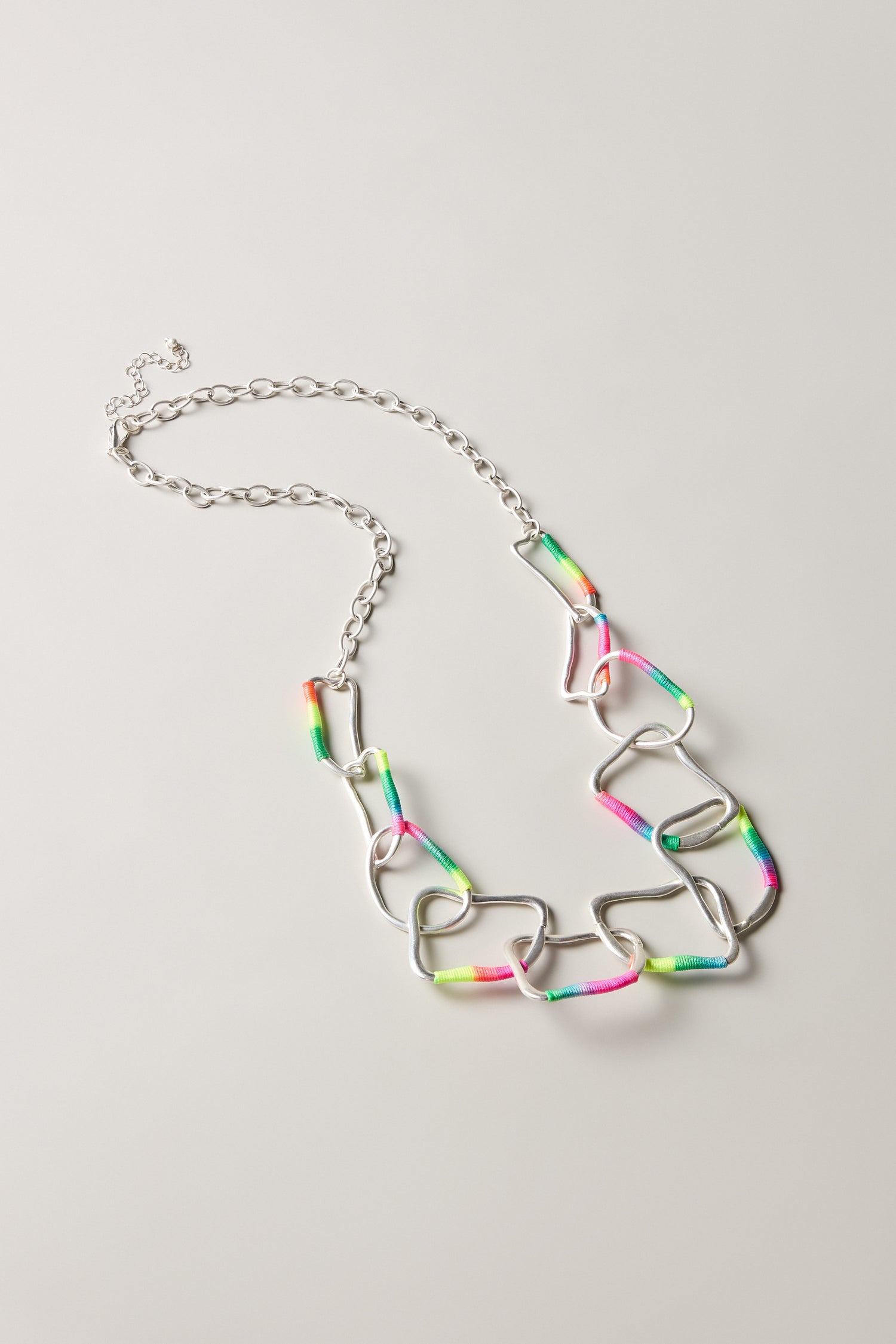 The Rainbow Detail Crushed Hoop Necklace features large, irregular silver links partially wrapped in neon pink, green, and yellow thread—perfect for colourful statement jewellery lovers—displayed on a light background.