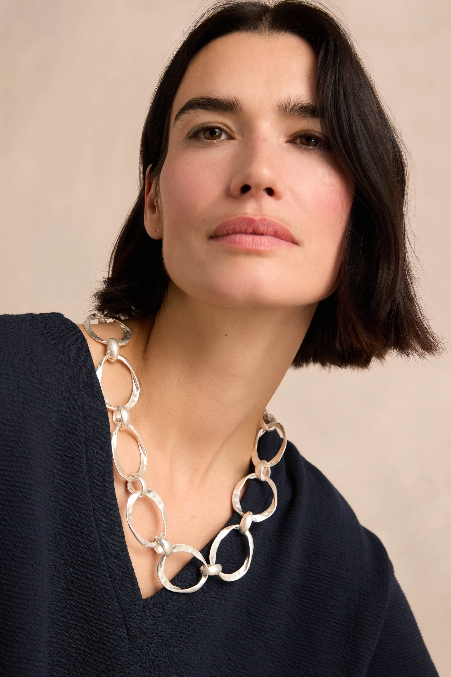 A woman with short dark hair wears a navy top and the Hammered Silver Hoop Necklace, featuring chunky silver-plated hammered hoop links, while looking slightly upward against a neutral background.