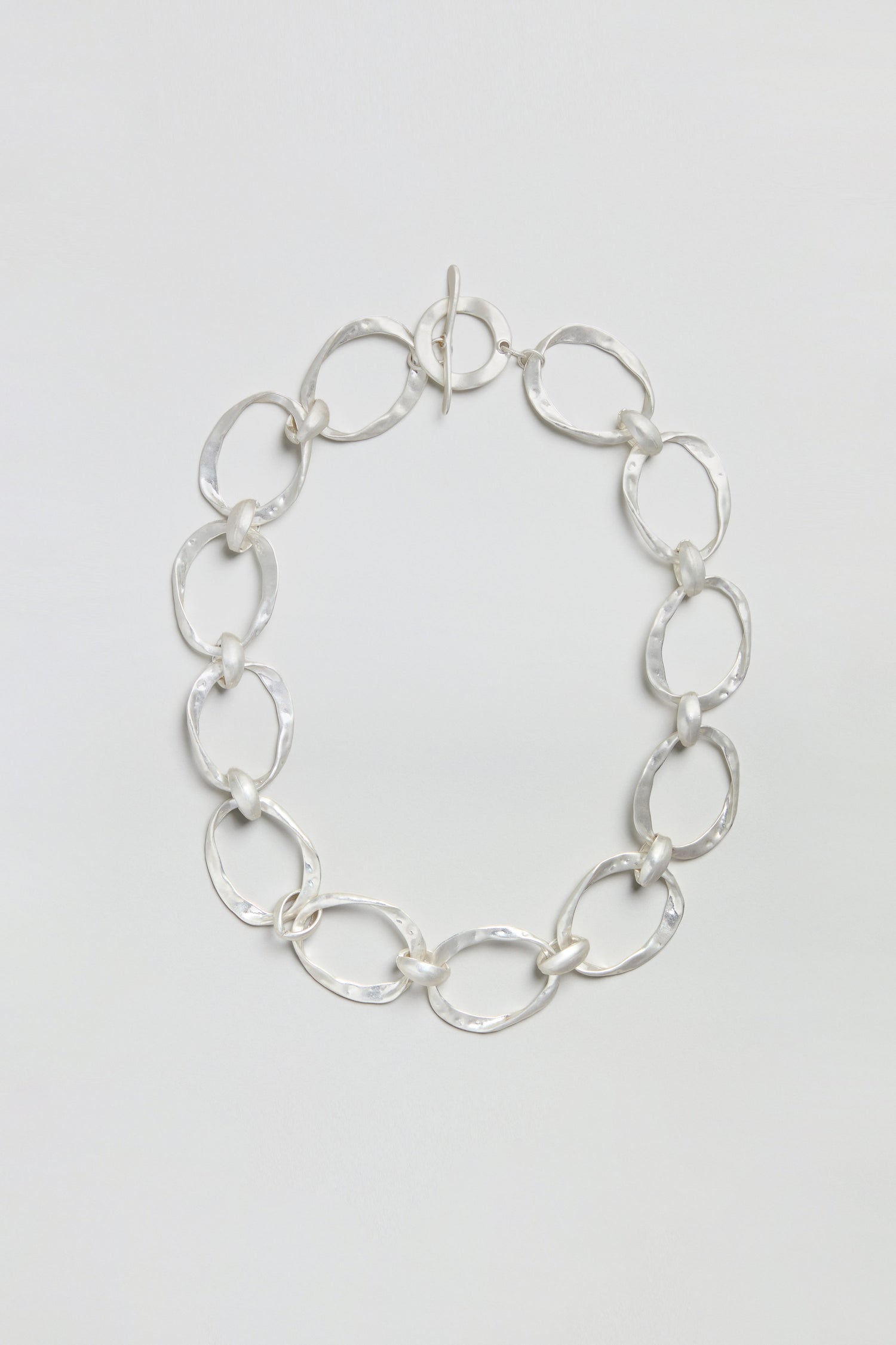 The Hammered Silver Hoop Necklace showcases large, irregular oval links with a hammered finish, joined by smaller round connectors, and is elegantly presented against a plain white background.