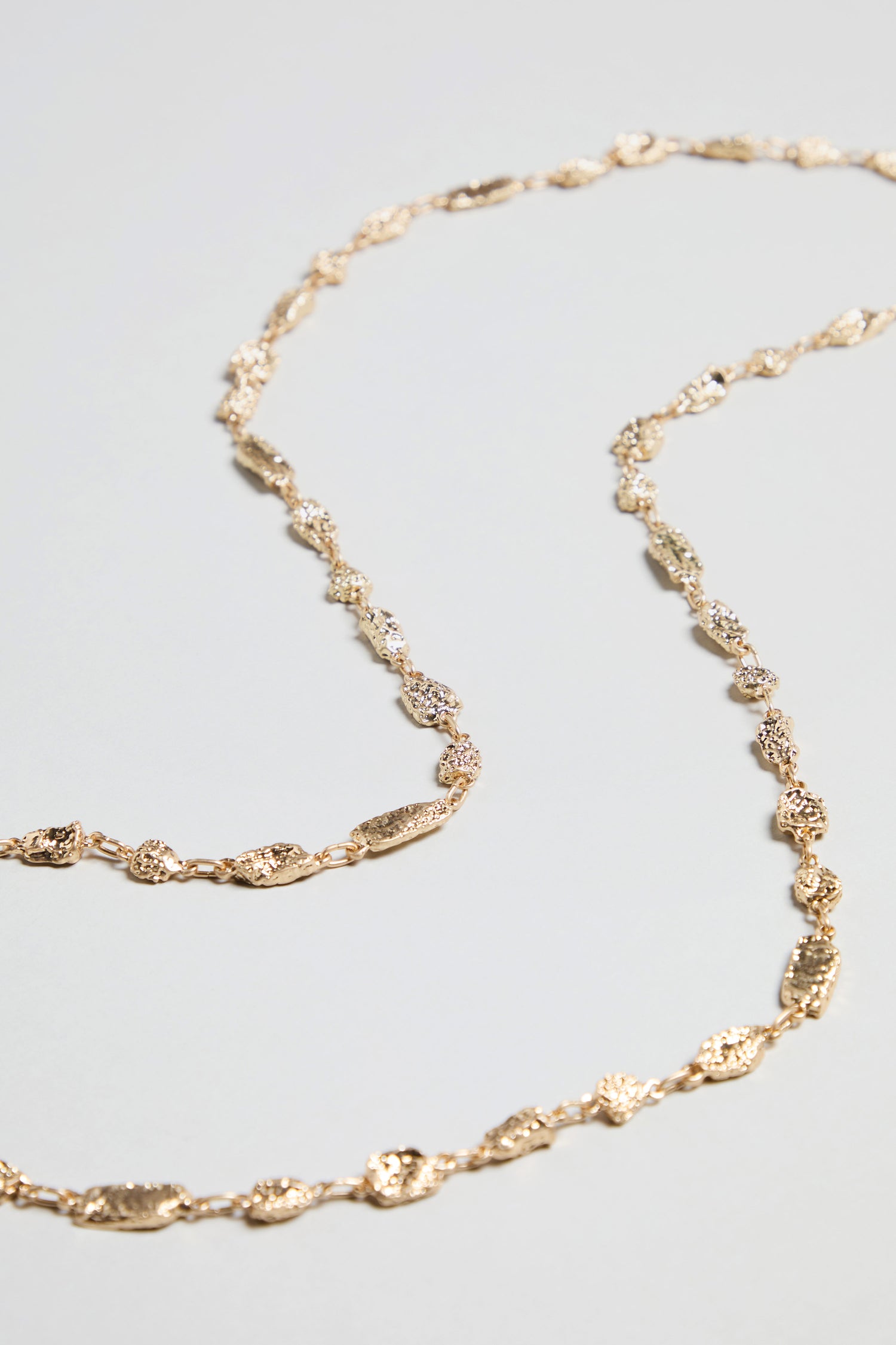 The Nugget Chain Necklace, featuring textured and irregularly shaped gold links, is displayed on a light grey surface.