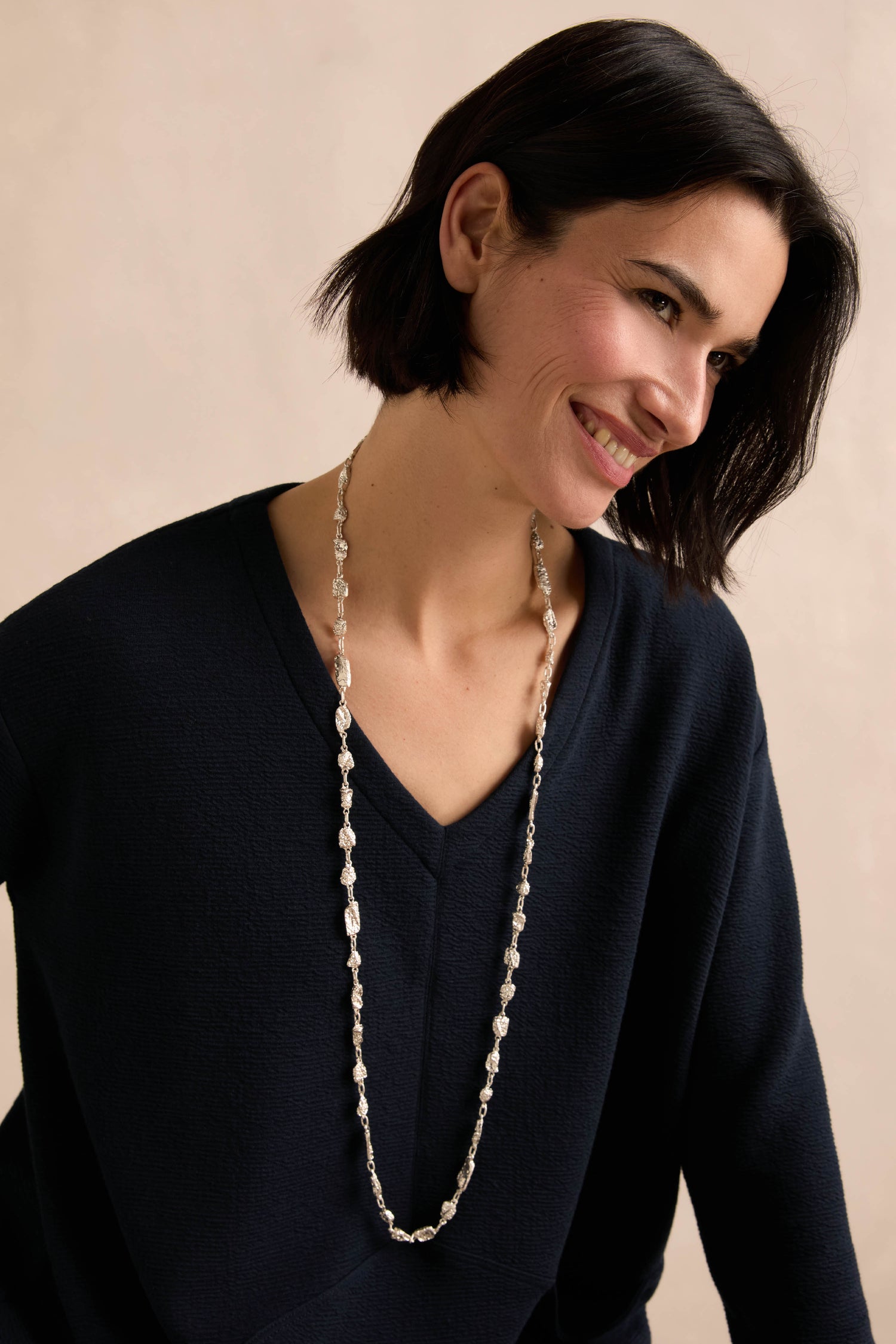 Wearing the Nugget Chain Necklace over a dark V-neck sweater, a woman with short dark hair smiles and looks slightly to the side against a neutral background.
