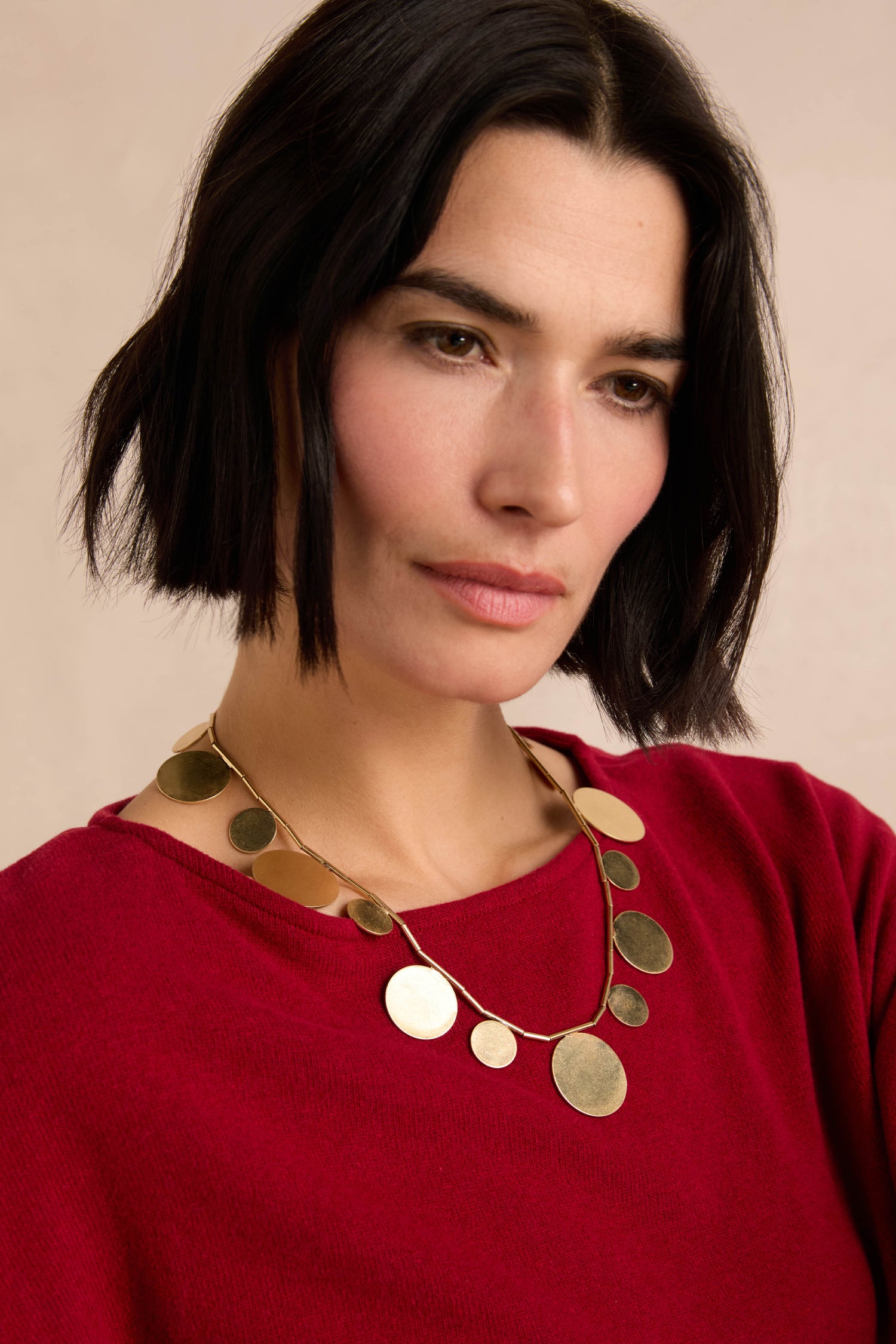 A woman with short black hair, dressed in a red sweater and wearing the Orbit Disc Necklace, looks slightly downward against a neutral background.