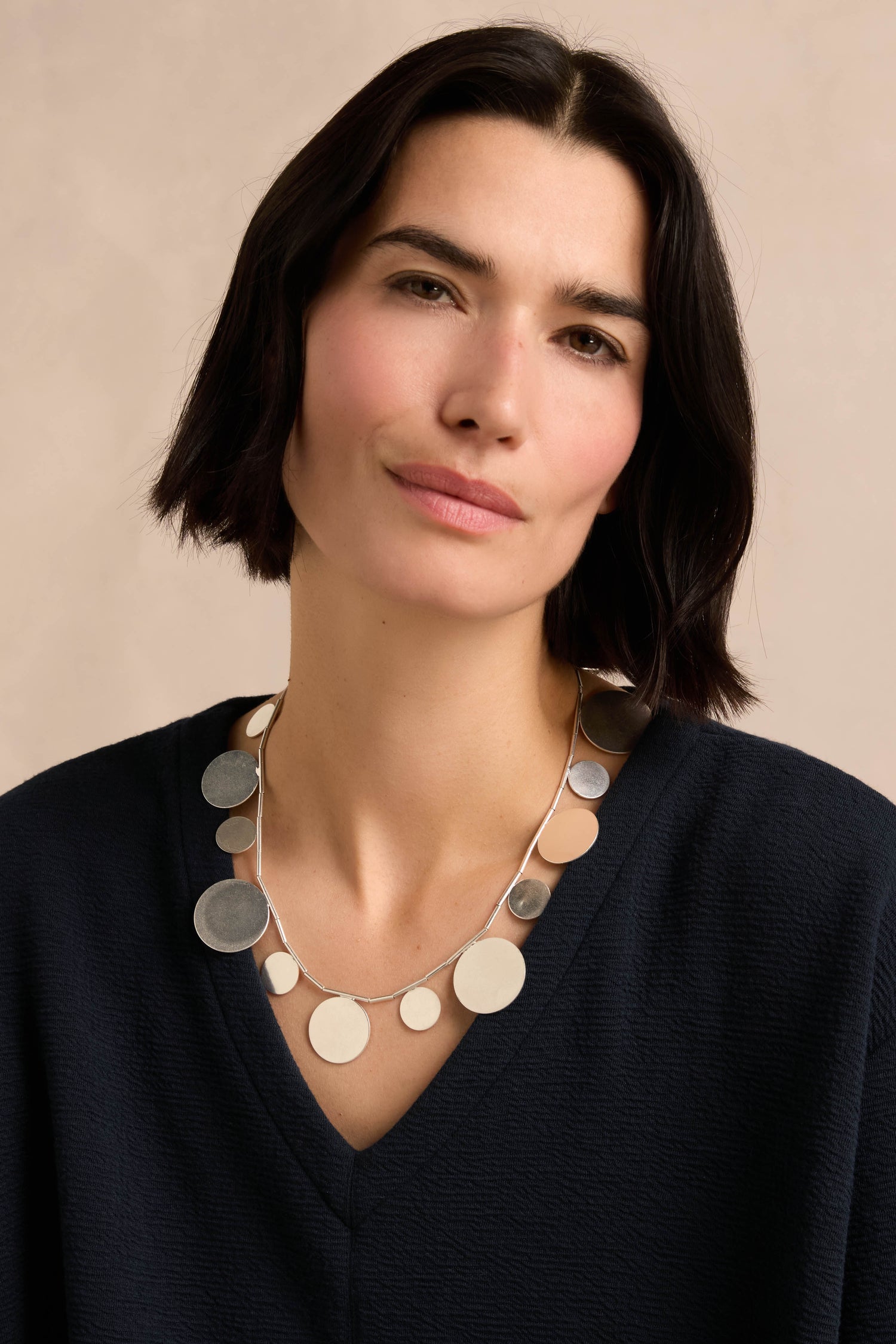 A woman with short dark hair wears a navy top and the Orbit Disc Necklace—a silver necklace featuring circular discs—as she poses against a neutral background.