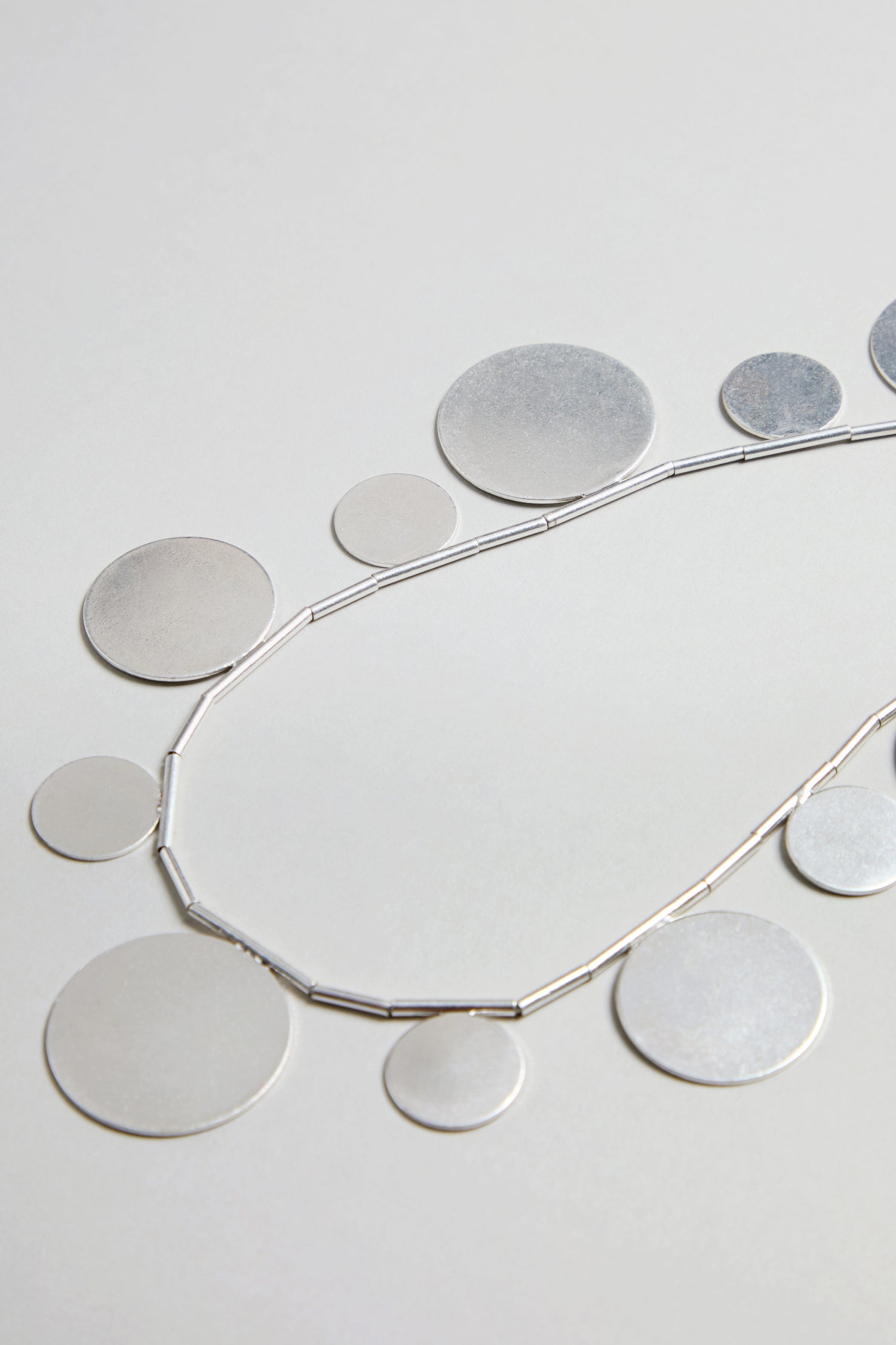 The Orbit Disc Necklace, featuring flat, round silver discs in assorted sizes, is displayed on a light grey background.