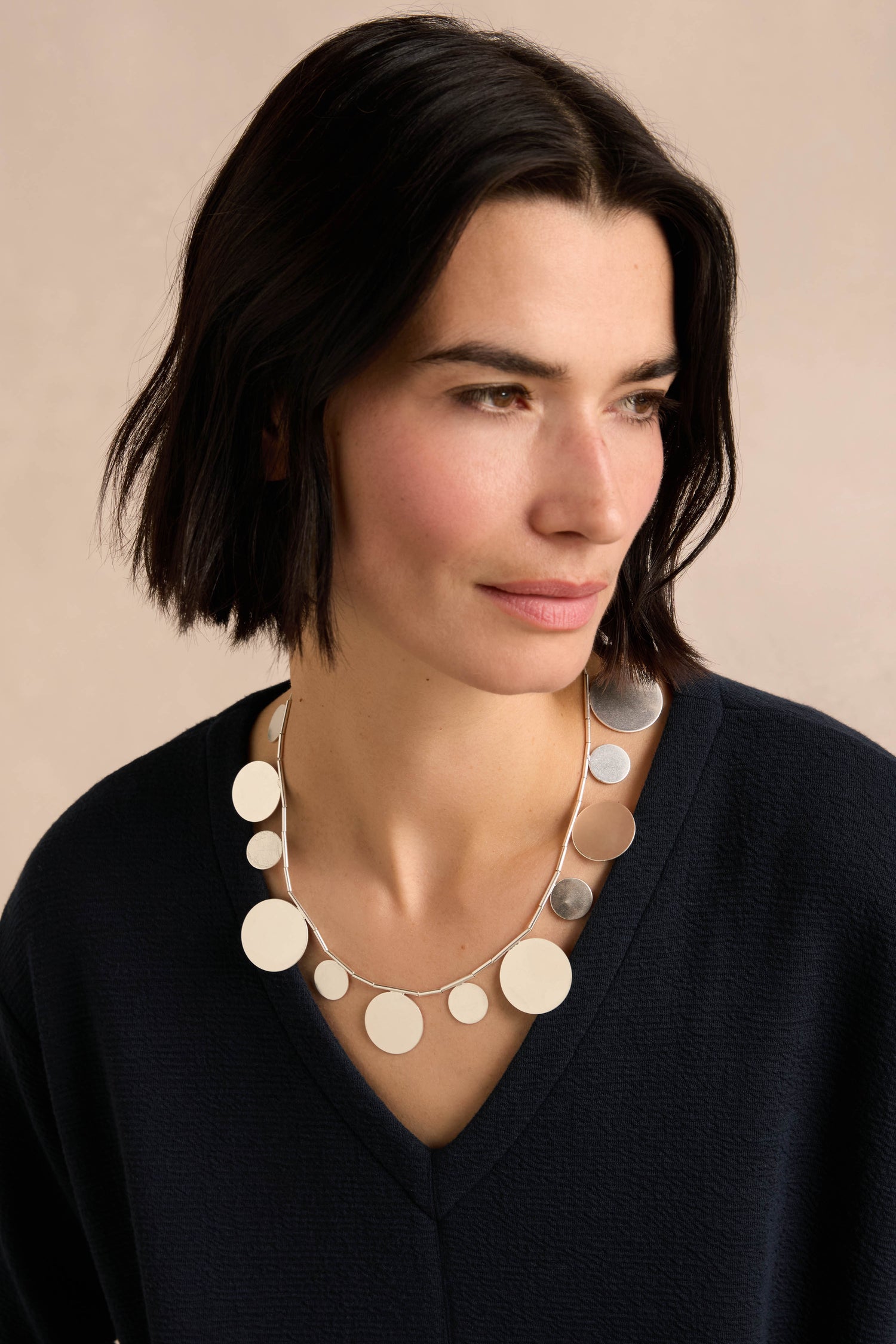 A woman with short dark hair wears a navy top and the Orbit Disc Necklace, featuring striking large, flat silver circular pendants, as she looks slightly to the side against a neutral background.