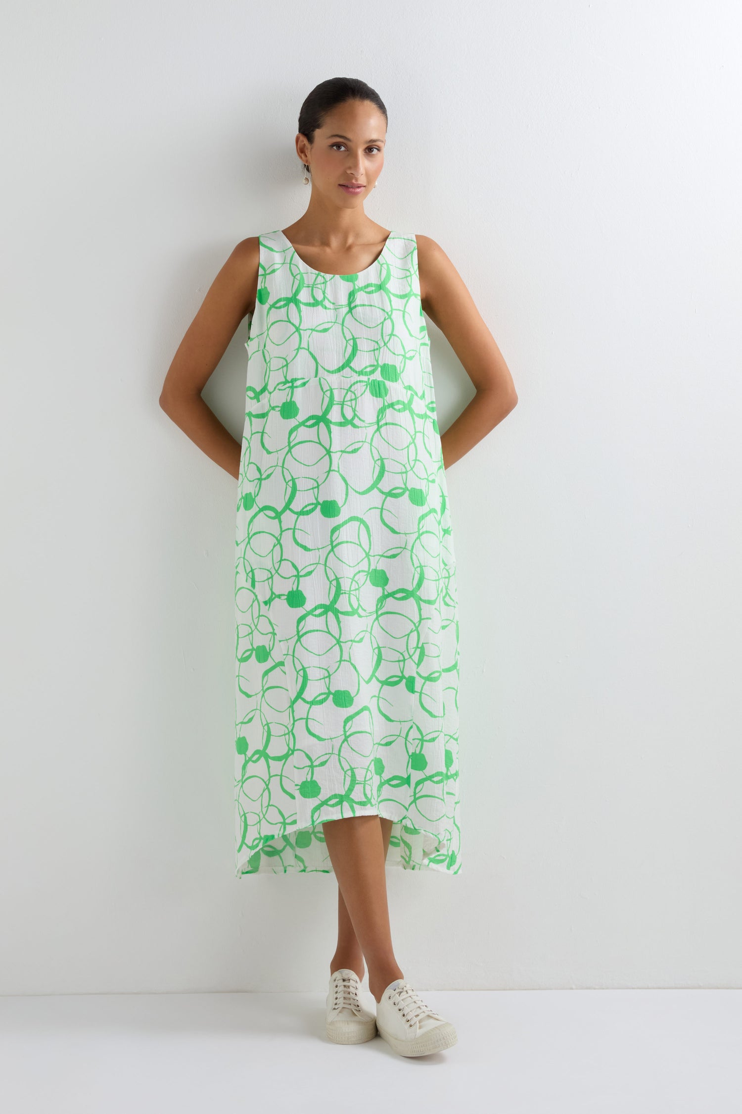 A woman stands against a white wall wearing the Swirl Print Bubble Dress, which features a bubble silhouette with a green swirl pattern, paired with white sneakers.
